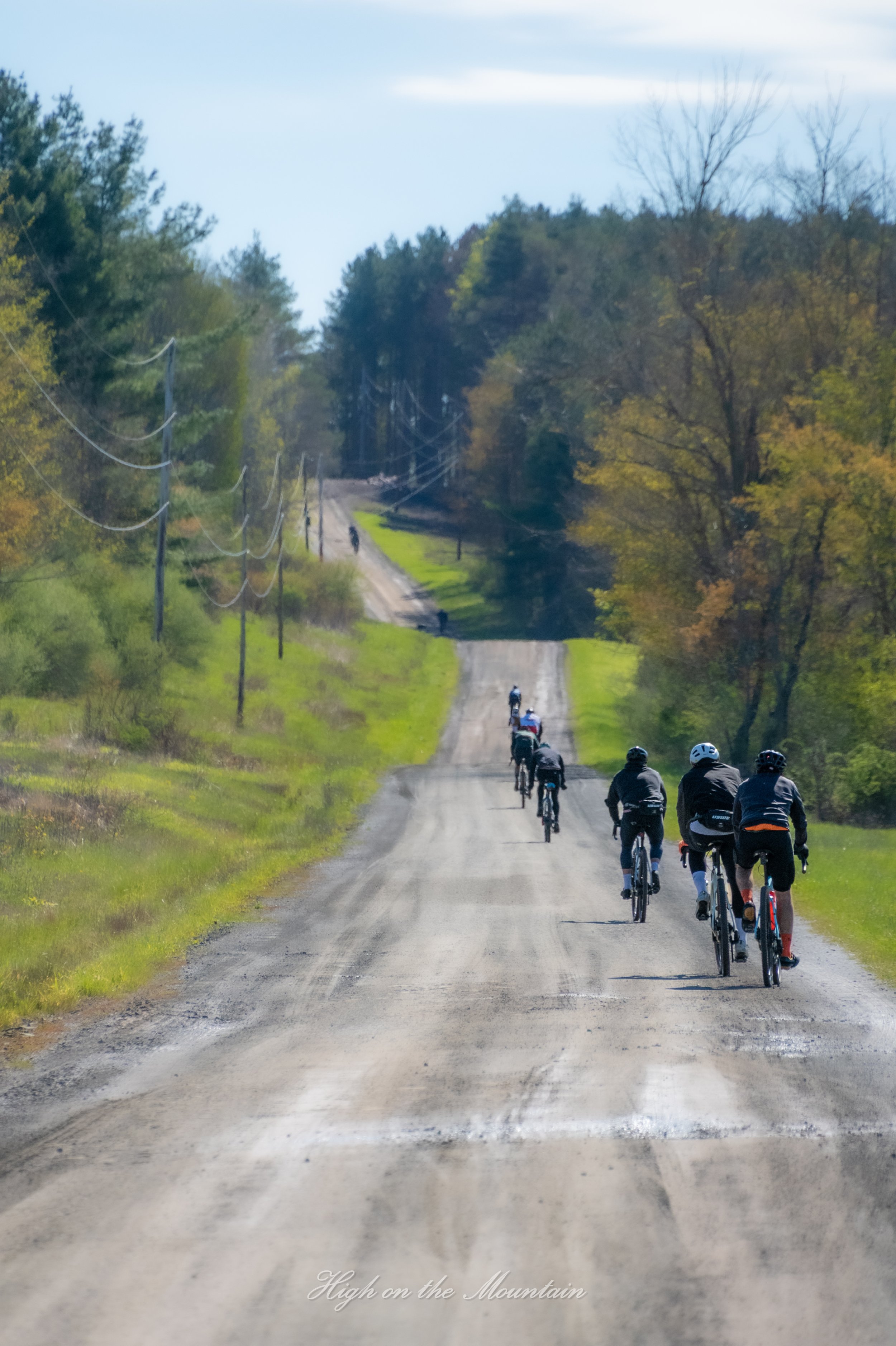 A group of cyclists riding on a dirt road through a scenic countryside with trees and utility poles lining the road. The sky is partly cloudy.