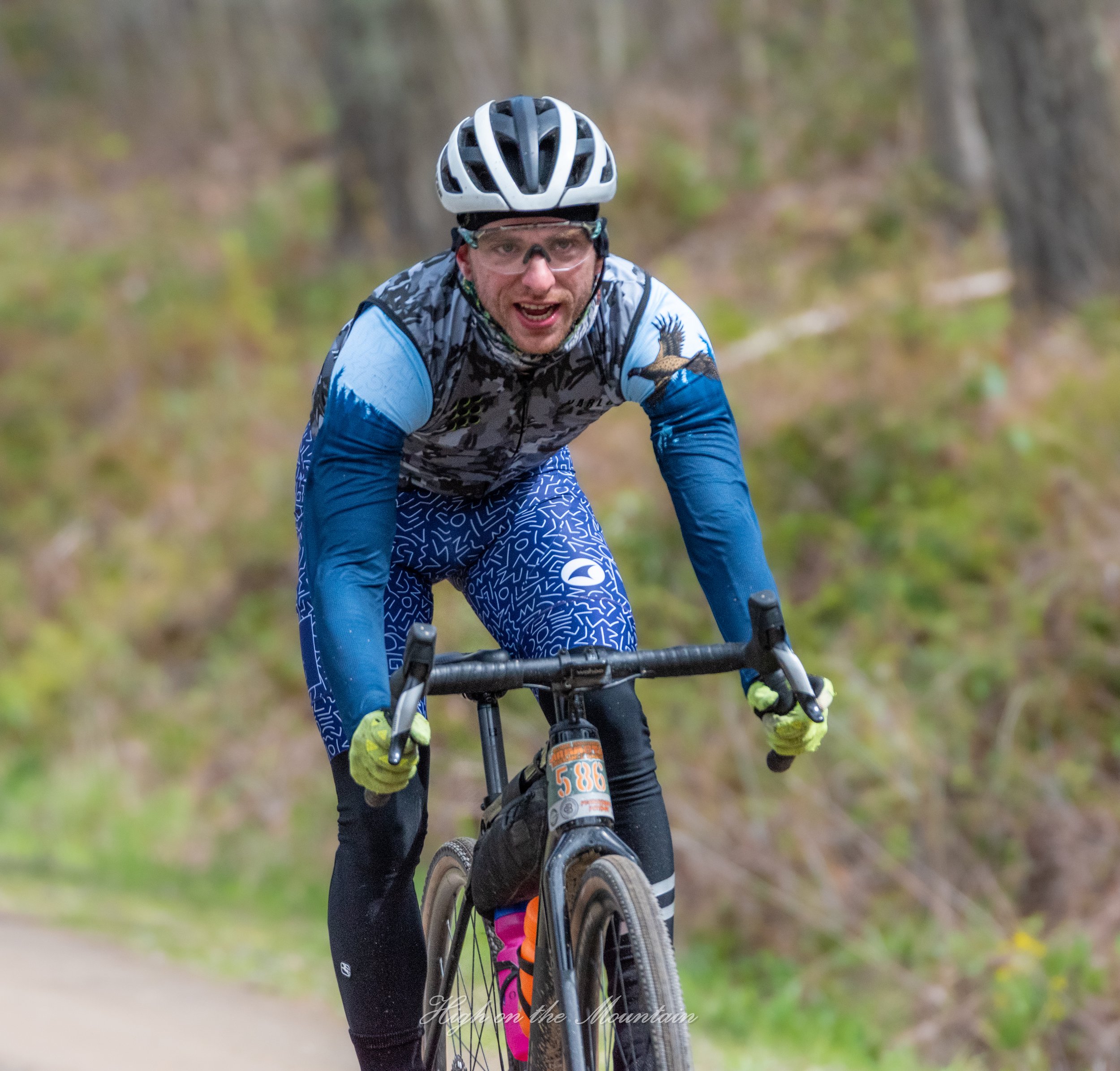 A person wearing a bike helmet, glasses, and a colorful cycling jersey is riding a mountain bike on a trail through a wooded area, with their face showing concentration and effort.