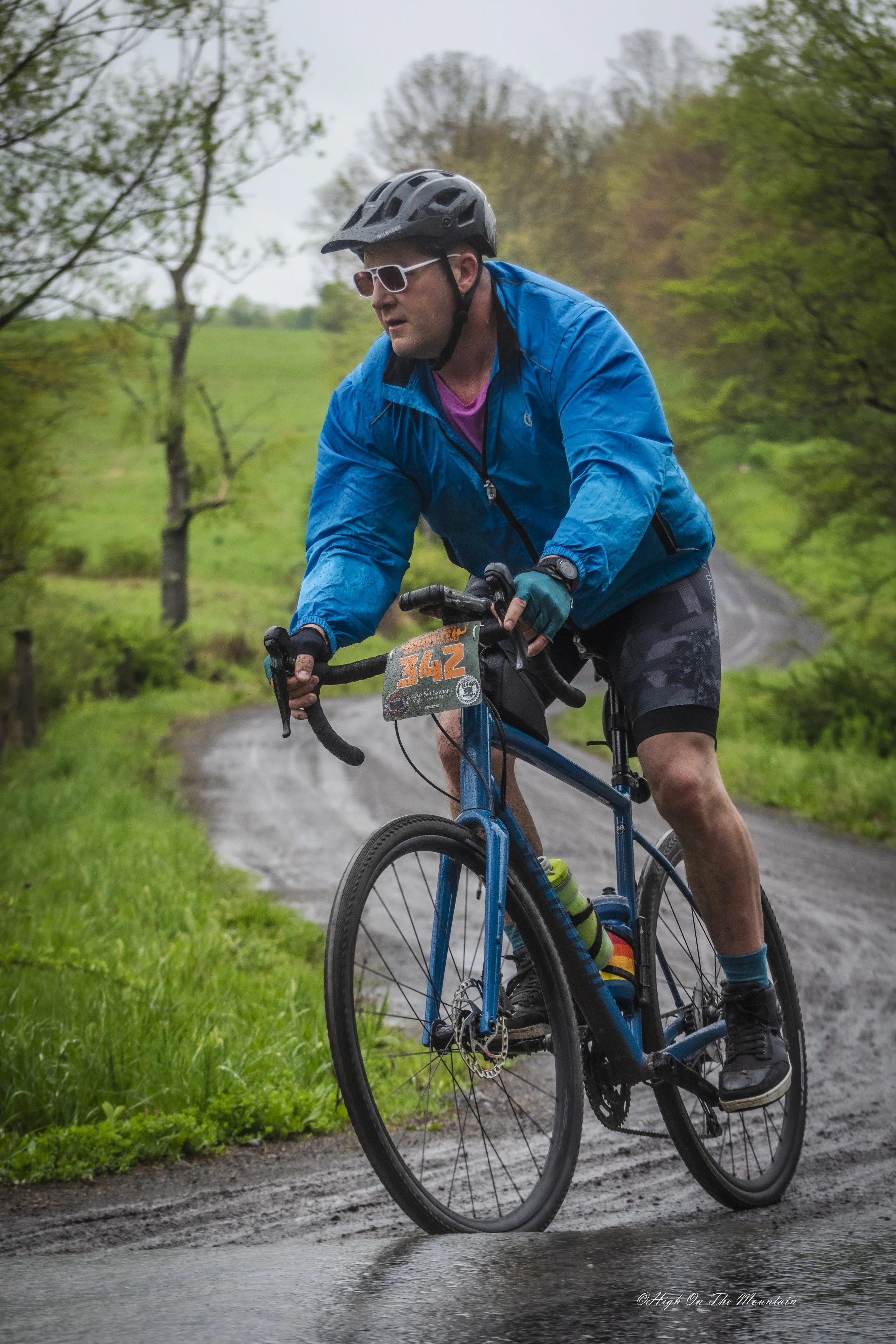 A man riding a mountain bike on a muddy trail in a green wooded area, wearing a black helmet, sunglasses, a blue jacket, and cycling shorts, with a number plate on his bike indicating he is participating in a race.