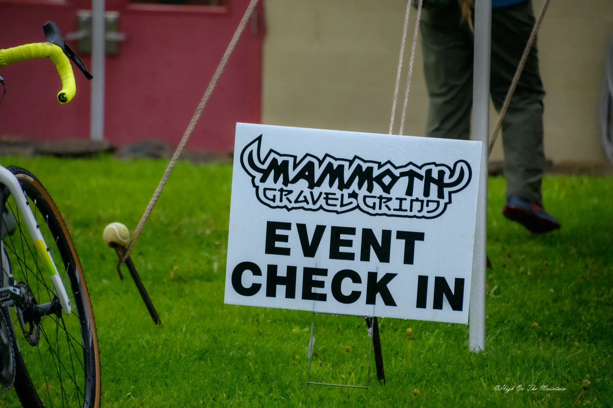 A bicycle tire and handlebar are visible on the left side, with a white sign in the center that reads 'TraveltheGrind Event Check In.' The background shows a person standing on a green grassy area, with part of a red wall and ropes to the right.