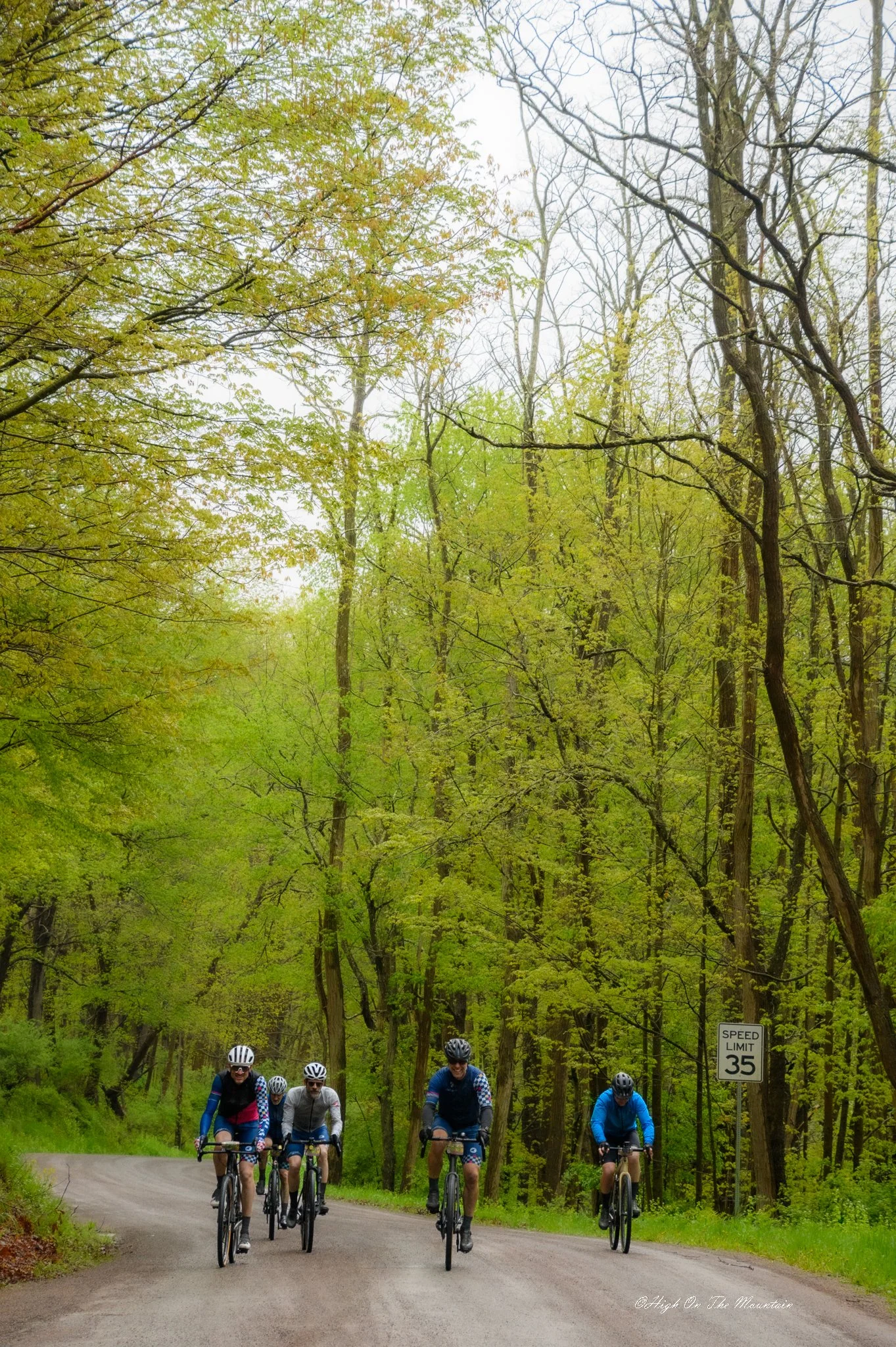 Group of cyclists riding bikes on a forested road with green leaves and a speed limit sign of 35 mph.