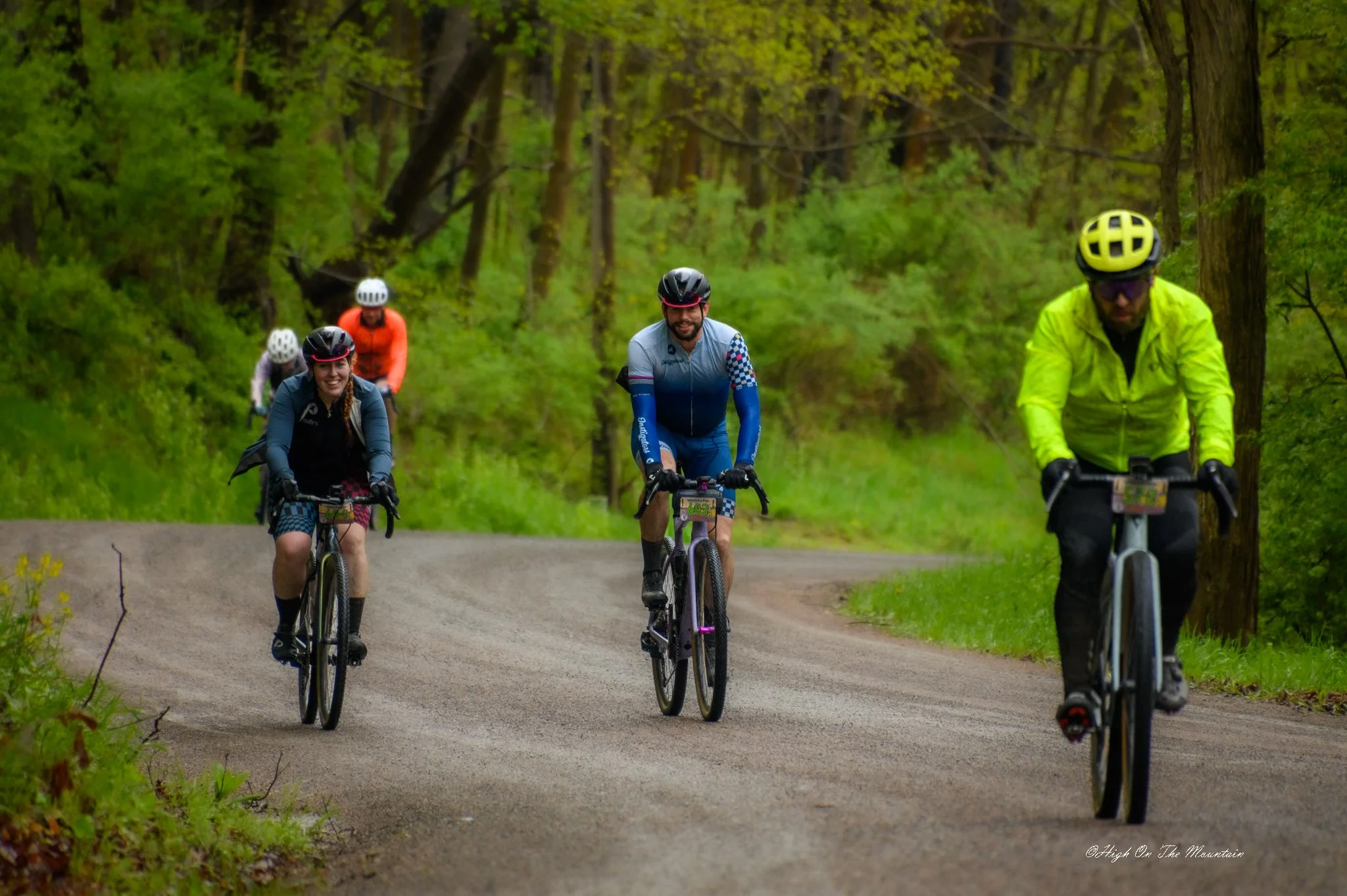 Group of five cyclists riding on a forest trail, surrounded by green trees and foliage.