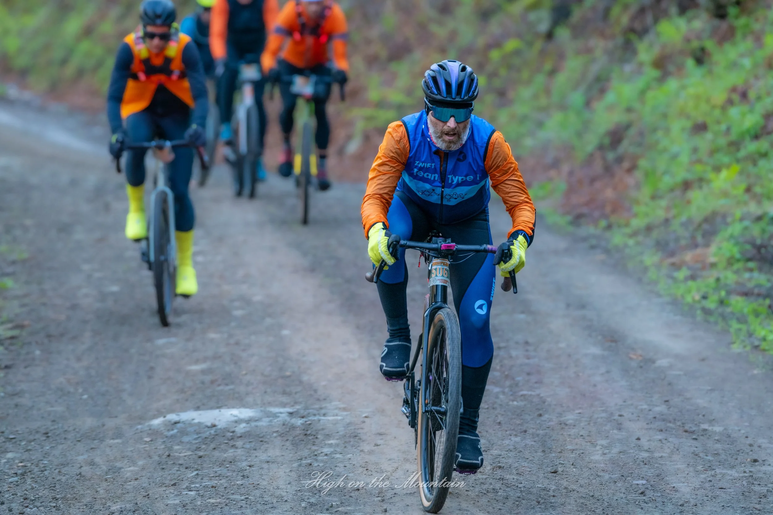 A group of mountain bikers riding on a dirt trail through a green forest. The front rider is an older man wearing a blue and orange outfit, black helmet, and sunglasses, with other riders following behind.