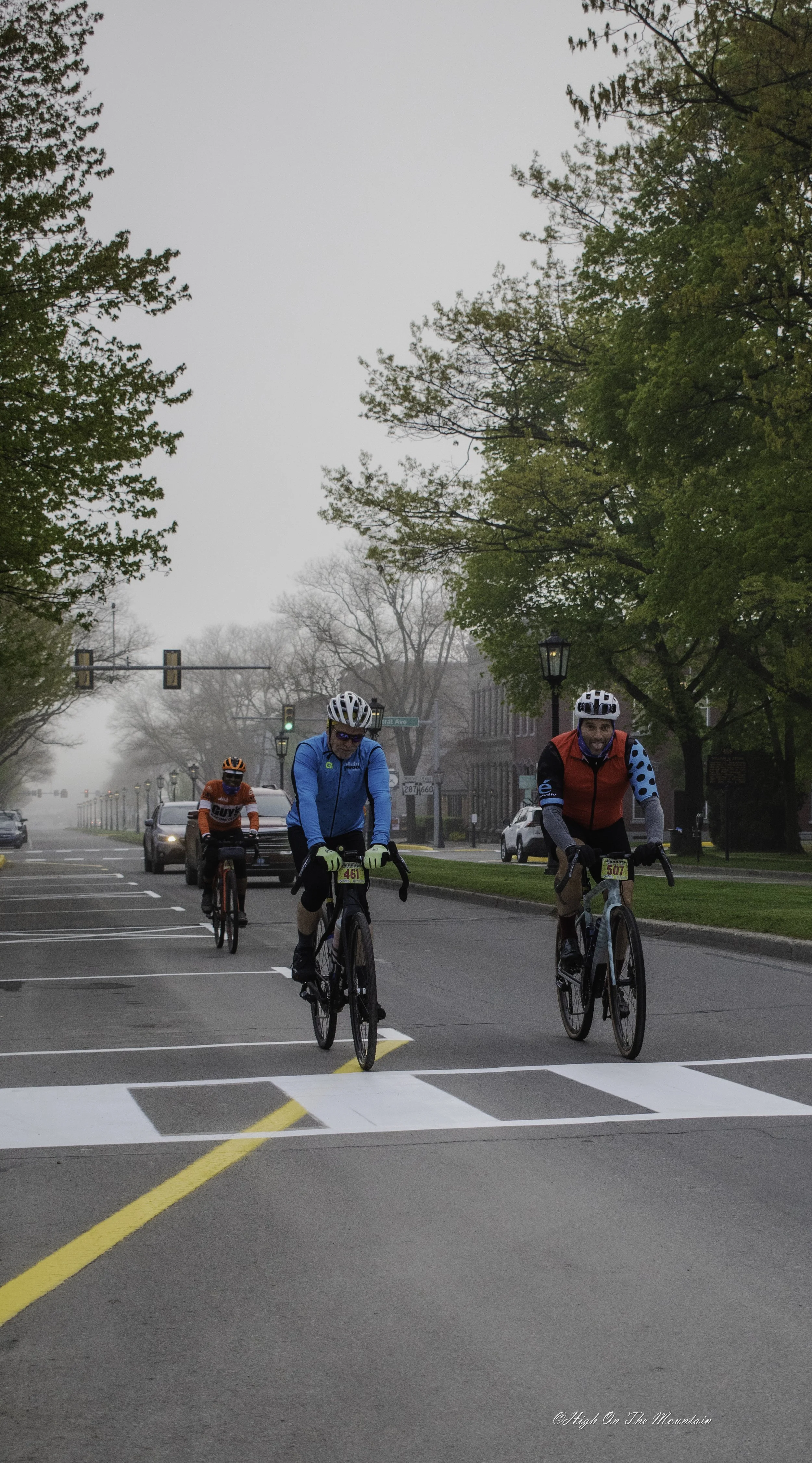 Three cyclists riding their bikes on a city street near a crosswalk with traffic lights, trees on both sides, and some cars parked and driving in the background on a foggy day.