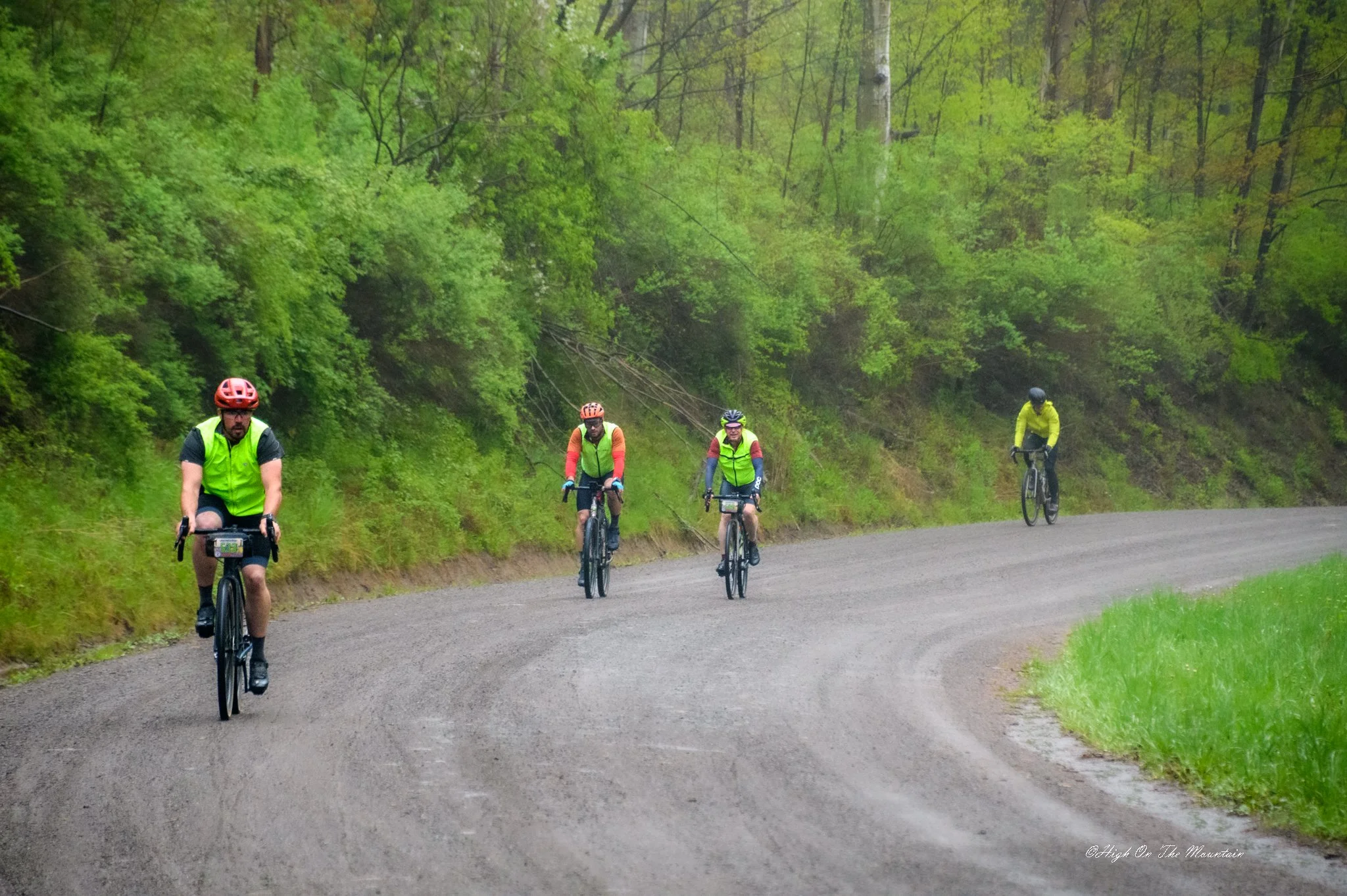 Four cyclists wearing helmets and reflective vests riding along a gravel trail through a lush green forest, with some trees visible in the background.