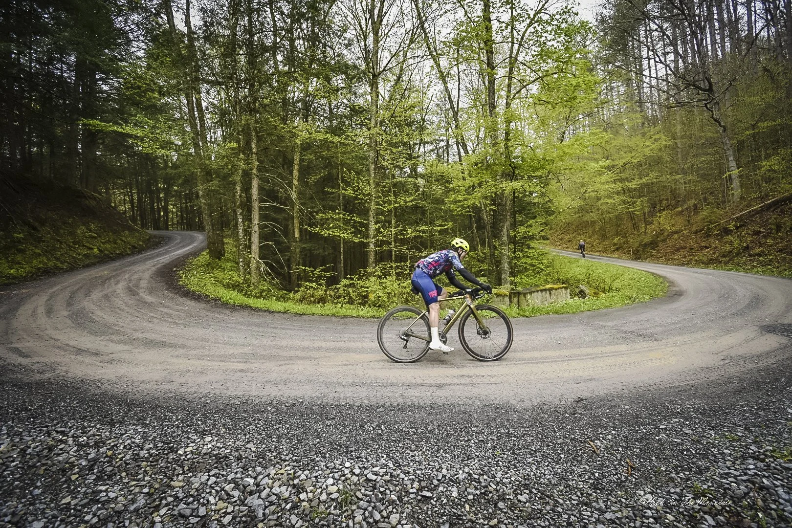 Cyclist riding a mountain bike on a winding gravel forest road, with another cyclist visible in the distance amidst dense green trees.