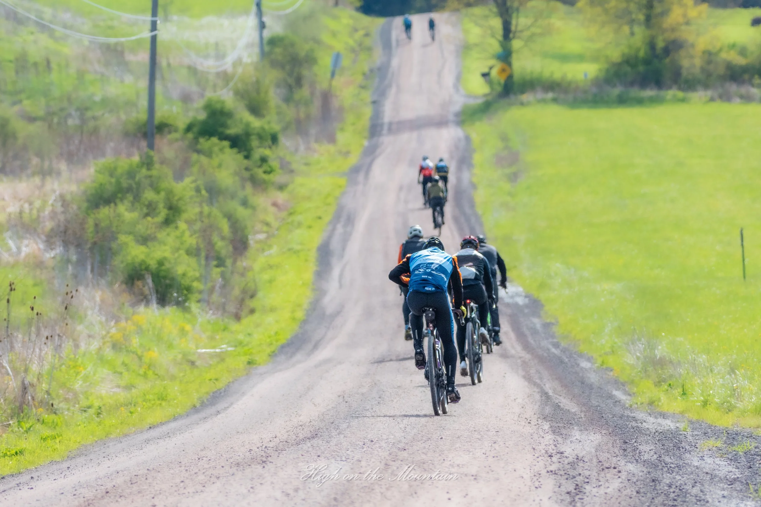A group of cyclists riding on a dirt road through a green, rural landscape with trees and power lines, with two people walking in the background.