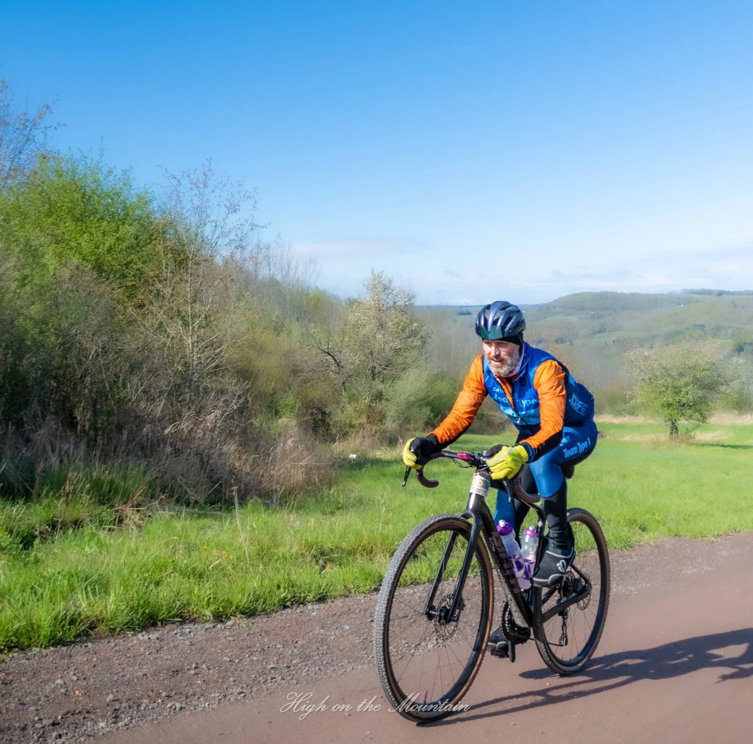 A person riding a mountain bike on a dirt trail through a scenic landscape with green hills and trees under a clear blue sky.