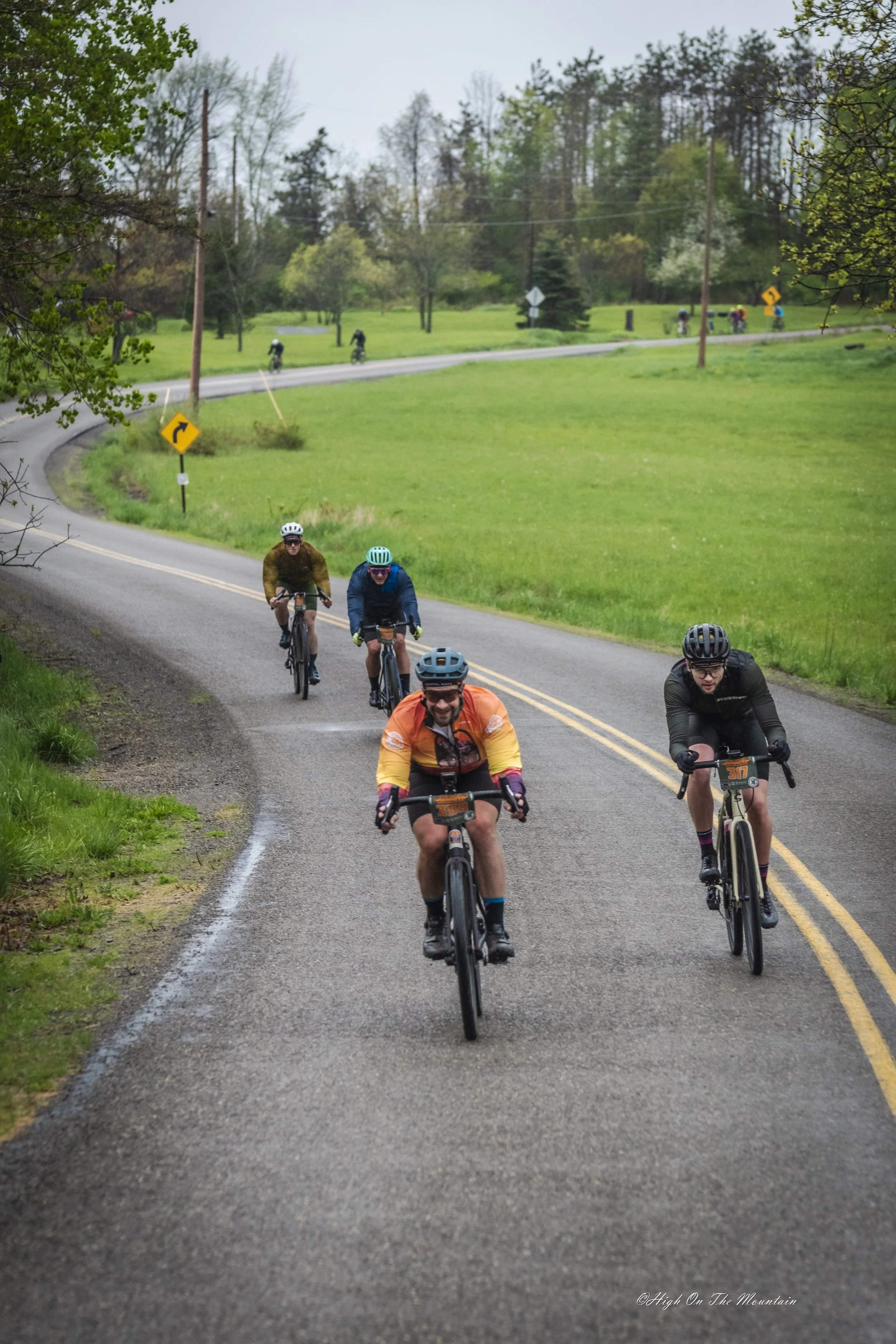 A group of cyclists riding on a winding road through a rural area with green grass and trees.