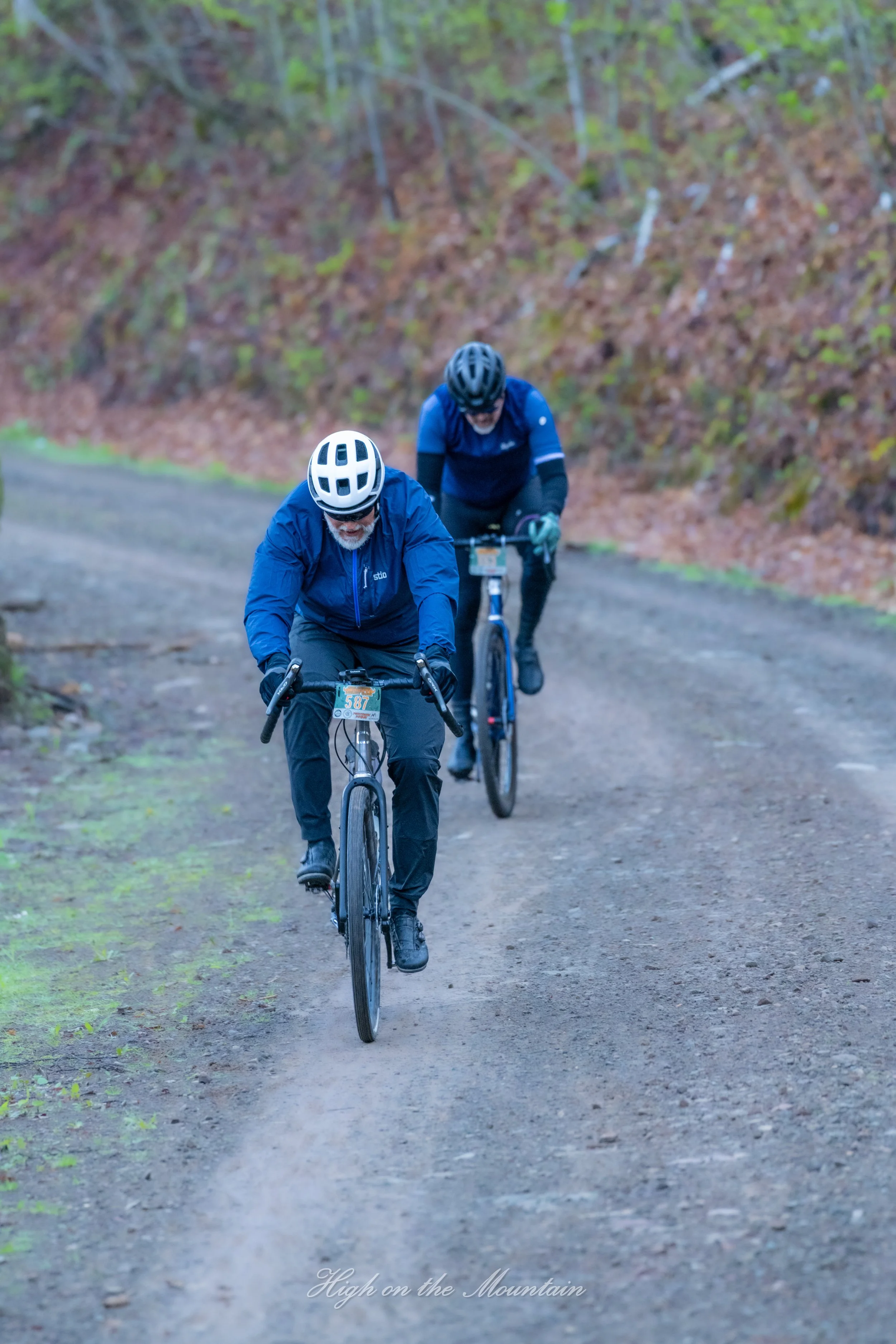 Two men riding mountain bikes on a dirt trail through a wooded area with brown and green leaves on the ground and trees in the background.