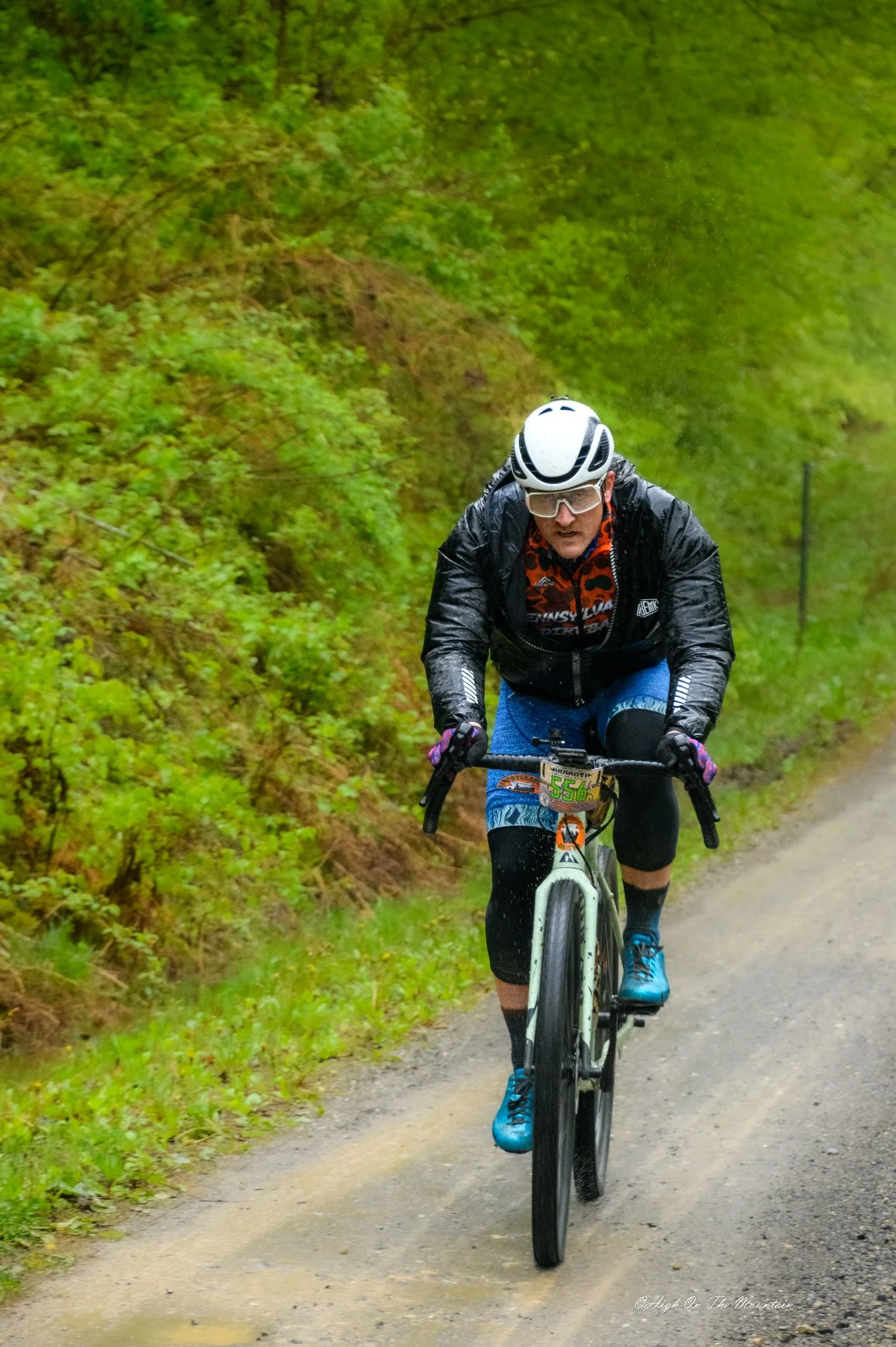 A person riding a mountain bike on a dirt trail through a green forested area. The rider is wearing a black rain jacket, blue shorts, a white helmet, and glasses.