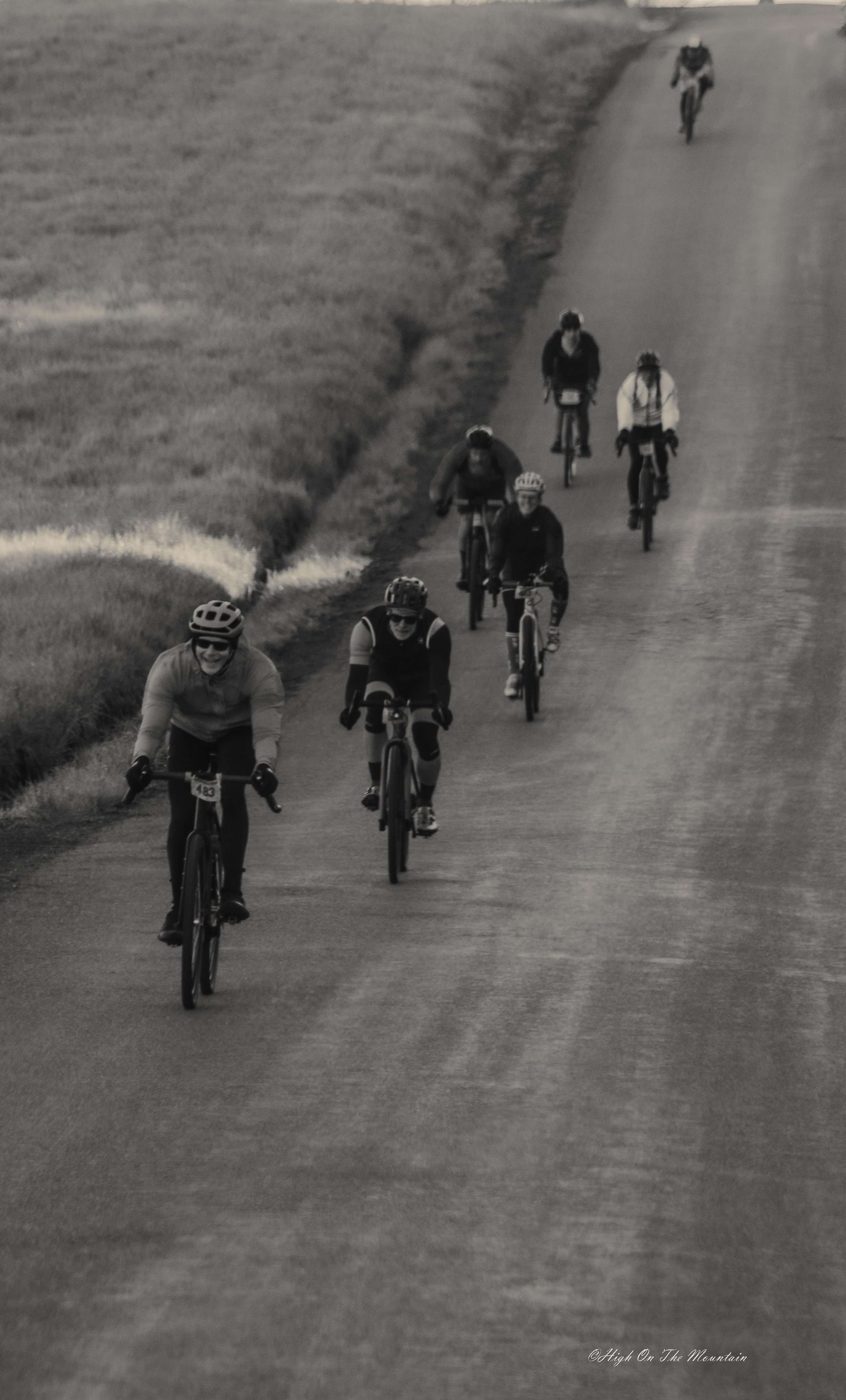 A group of people riding bikes along a rural, curved road with grassy fields on either side.