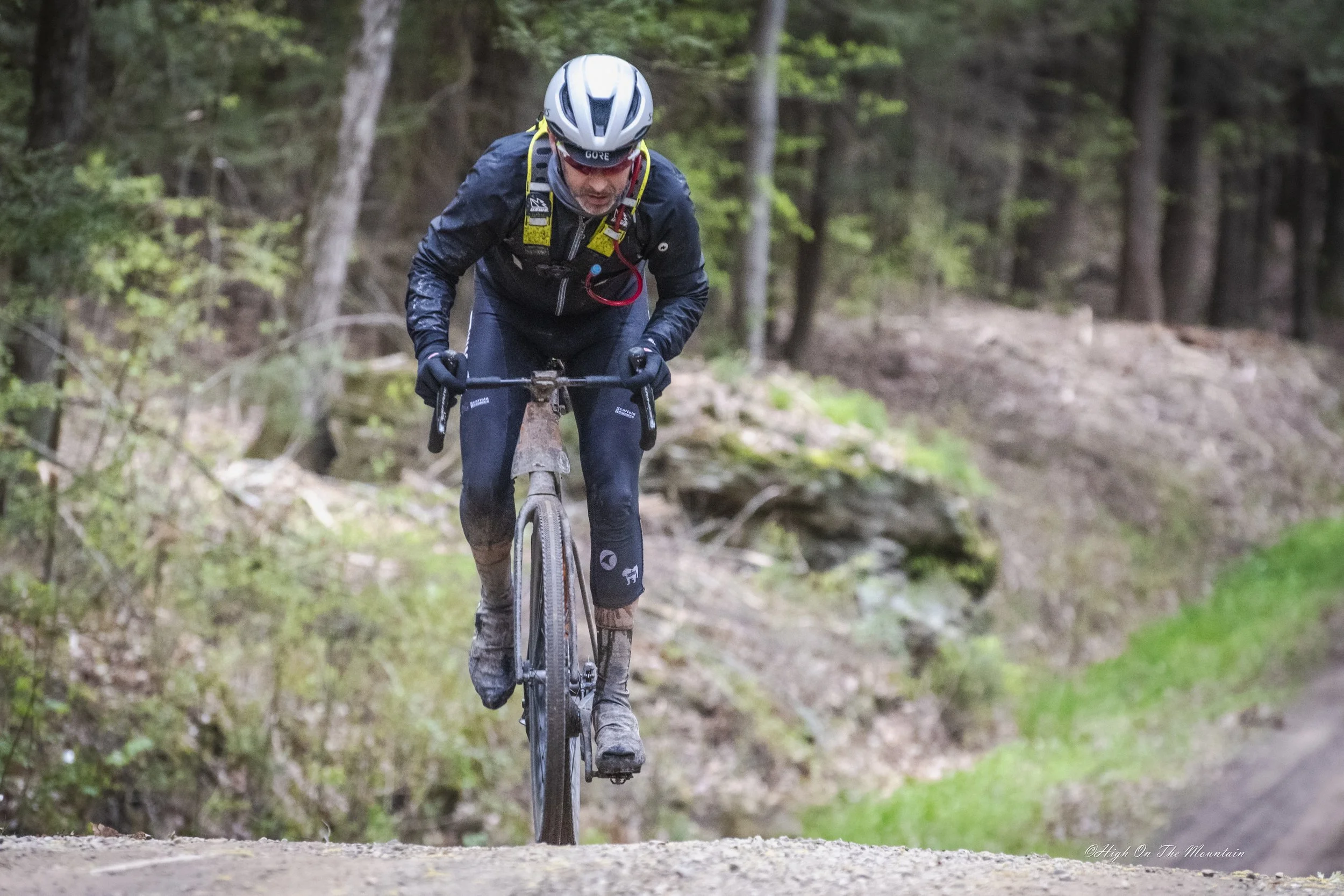 A man mountain biking on a forest trail, wearing a helmet, black jacket, and gloves, with a backpack and riding gear.