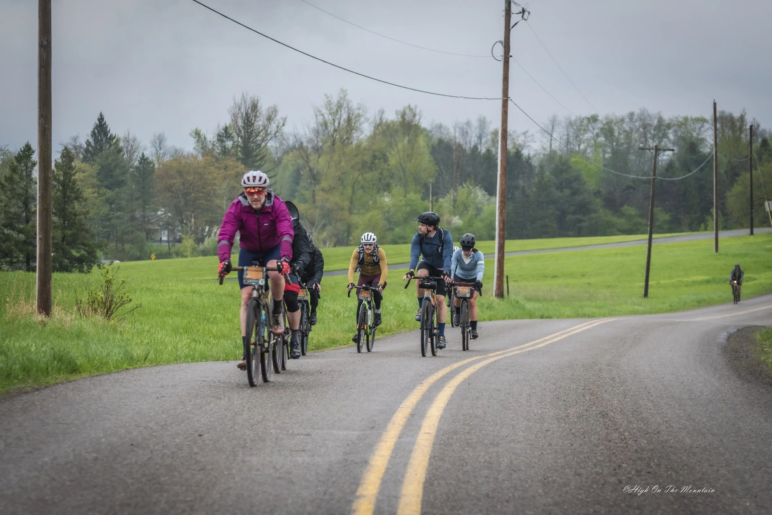 Group of six cyclists riding on a rural road with green fields and trees, overcast sky, wearing helmets and outdoor gear.