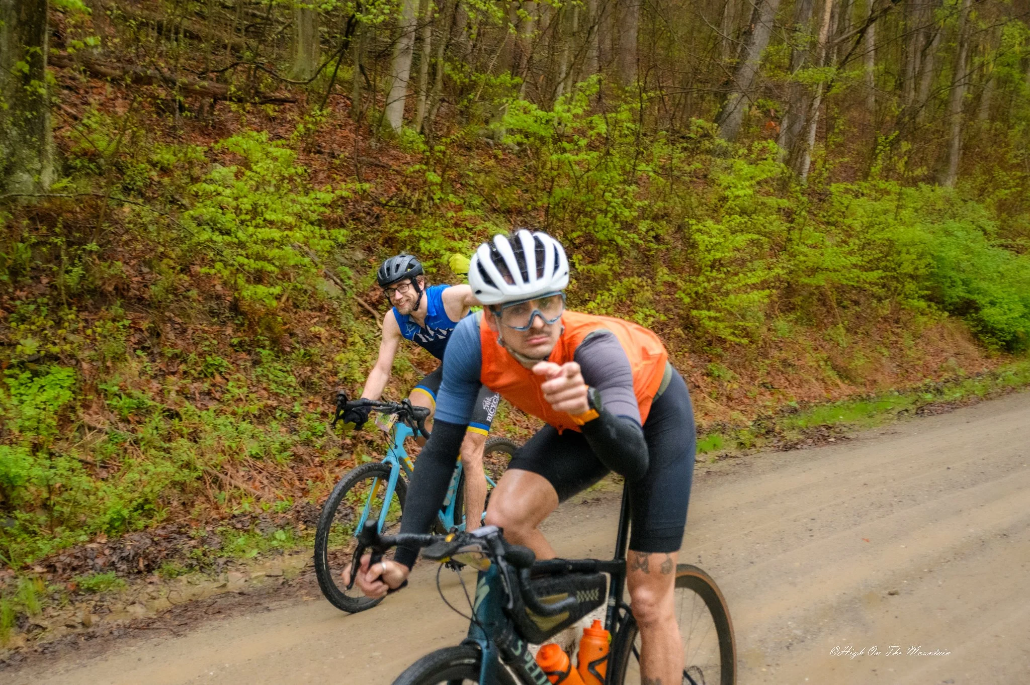 Two men riding mountain bikes on a trail in a wooded area, wearing helmets and athletic clothing, with green foliage and trees in the background.