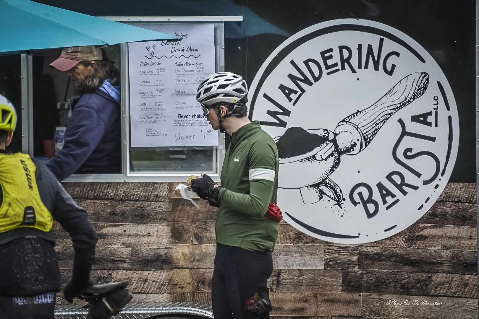 Cyclists in front of a seafood bar with a menu, near a seafood restaurant called WANDERING STARFISH BAR. One cyclist is eating a snack, while others are in cycling gear with helmets.