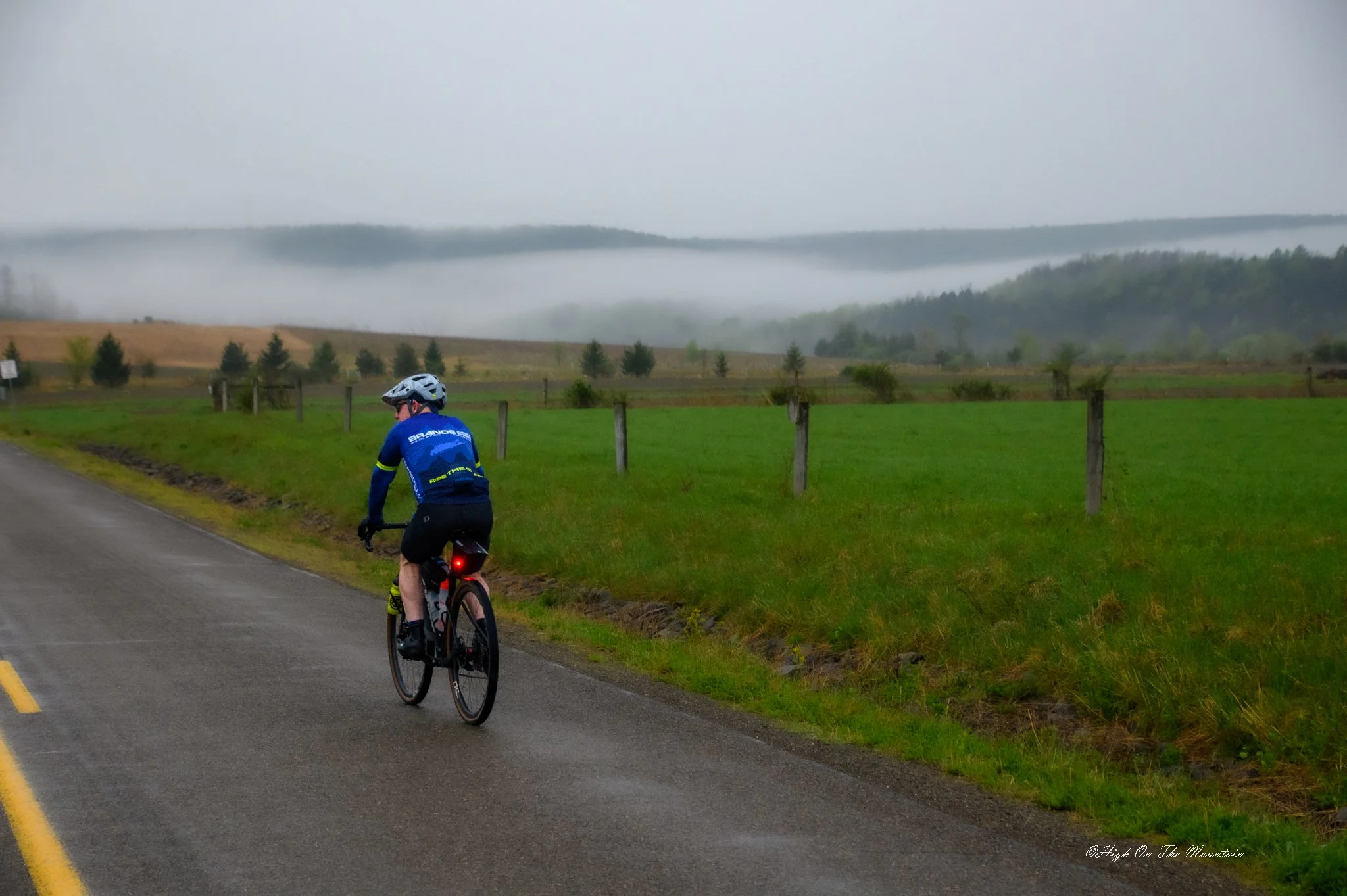 A person riding a bicycle along a paved road in a rural area with green grass, trees, fog, and hills in the background on an overcast day.