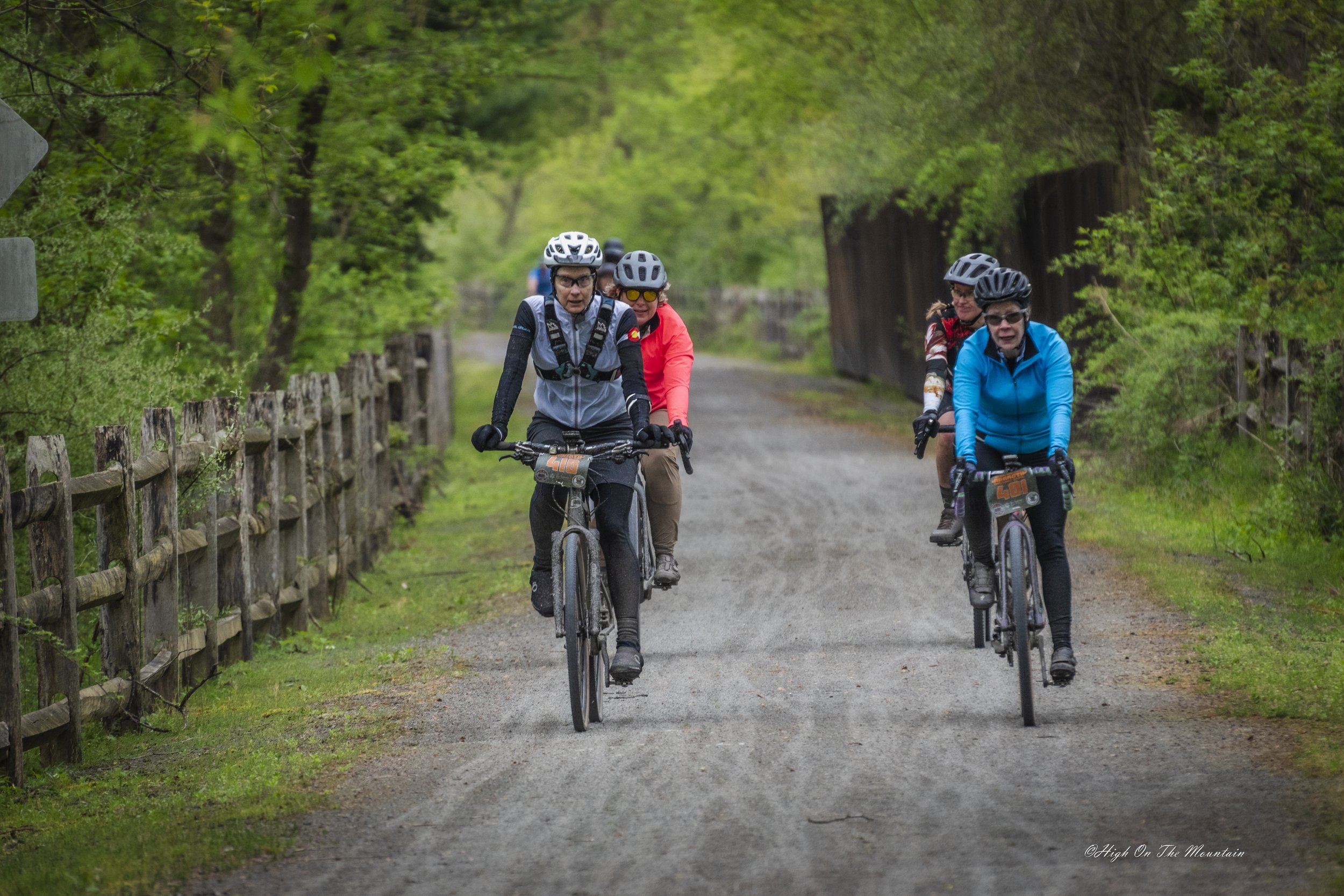 Four people riding mountain bikes on a dirt trail through a green, wooded area. They are wearing helmets and outdoor gear, with two women in the front and two men behind.