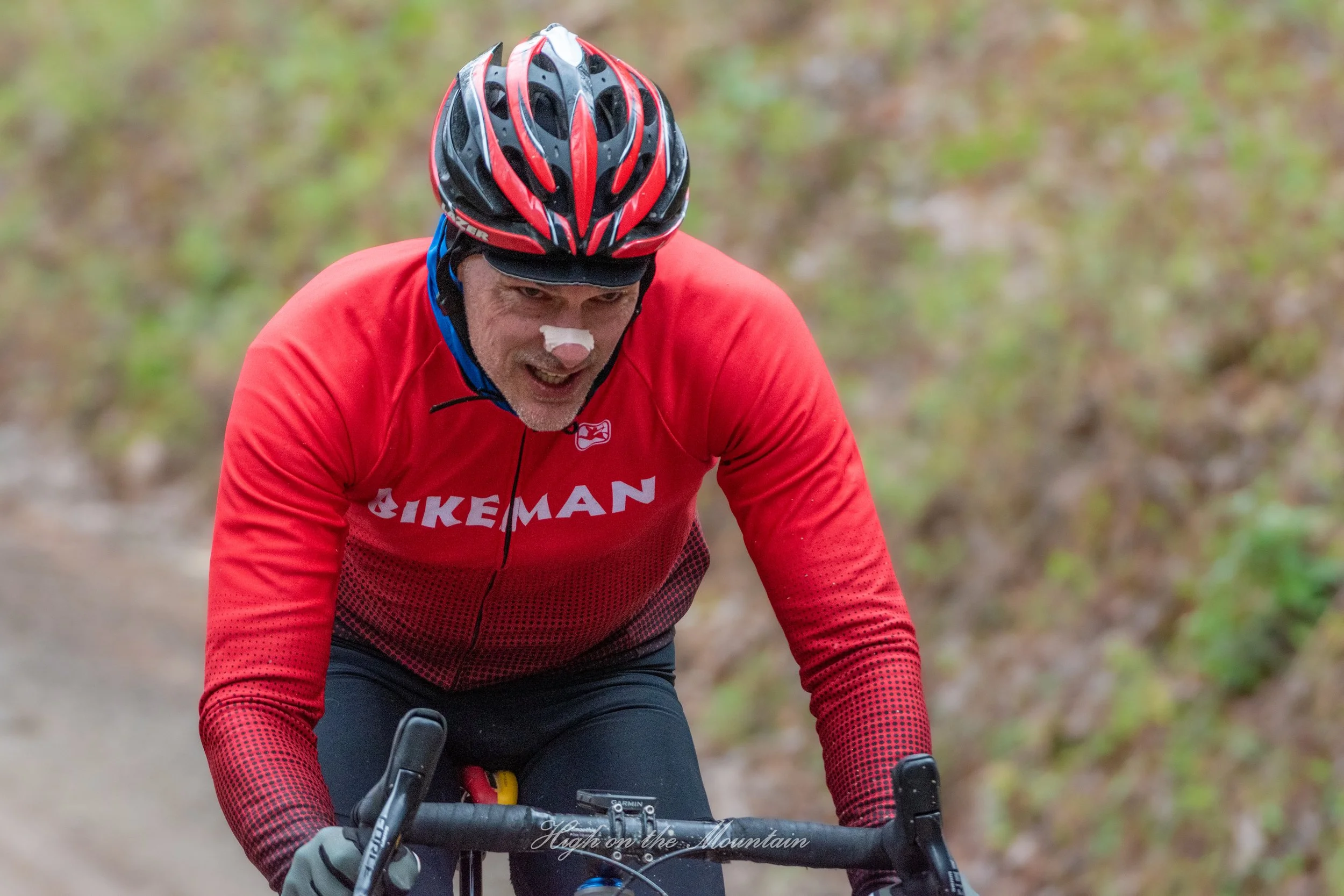 A man wearing a red cycling jersey, black helmet, and gray gloves is riding a mountain bike on a dirt trail surrounded by trees and foliage.