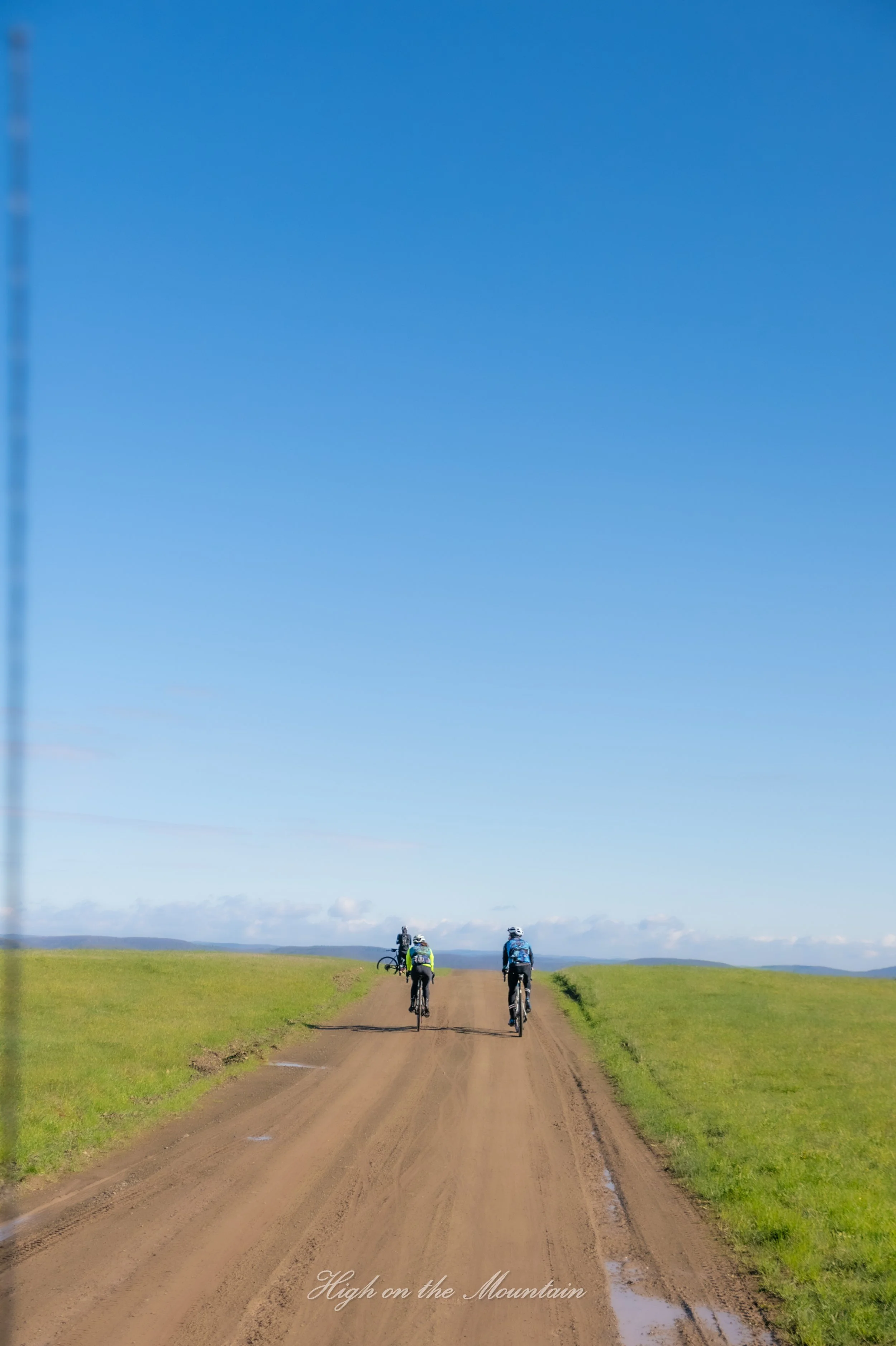 Three cyclists riding on a dirt path through green fields under a bright blue sky with scattered clouds.