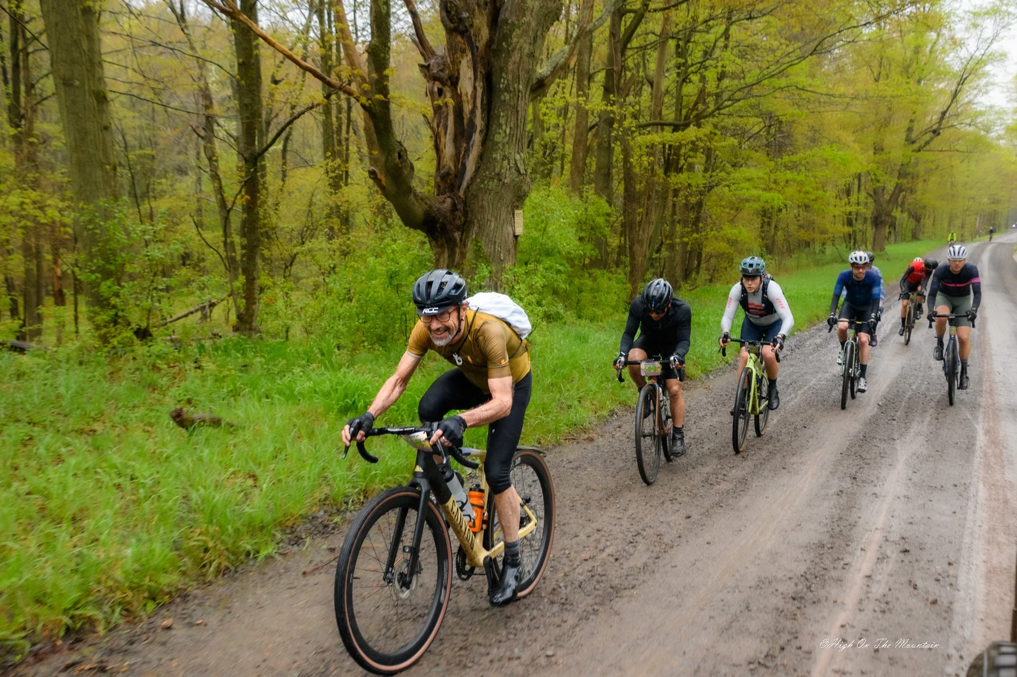 A group of mountain bikers riding on a dirt trail through a green, wooded forest during the daytime