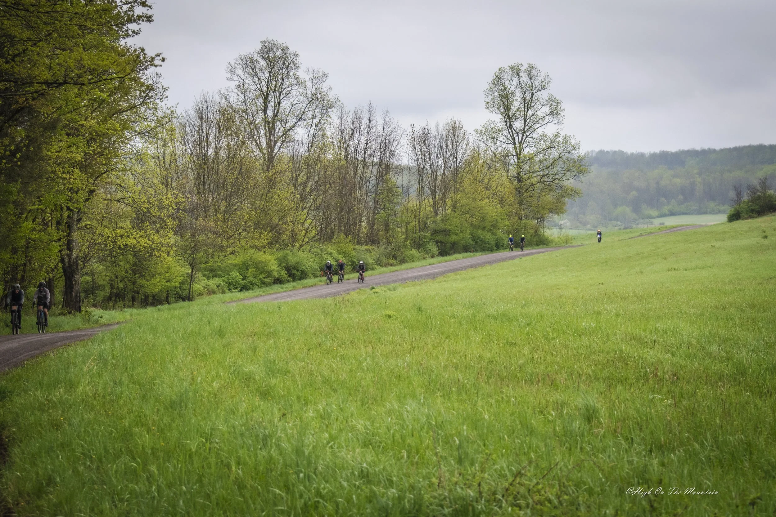 A group of cyclists riding on a dirt path through a green, hilly landscape with trees on both sides and a cloudy sky overhead.