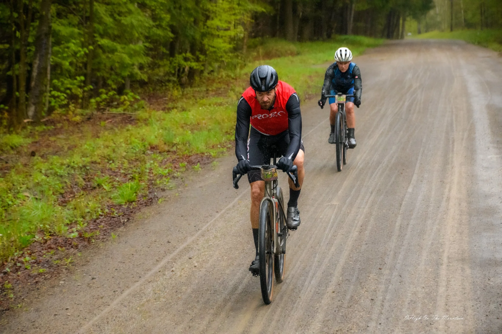 Two cyclists riding on a dirt trail through a green, wooded area with trees and grass on either side of the path.