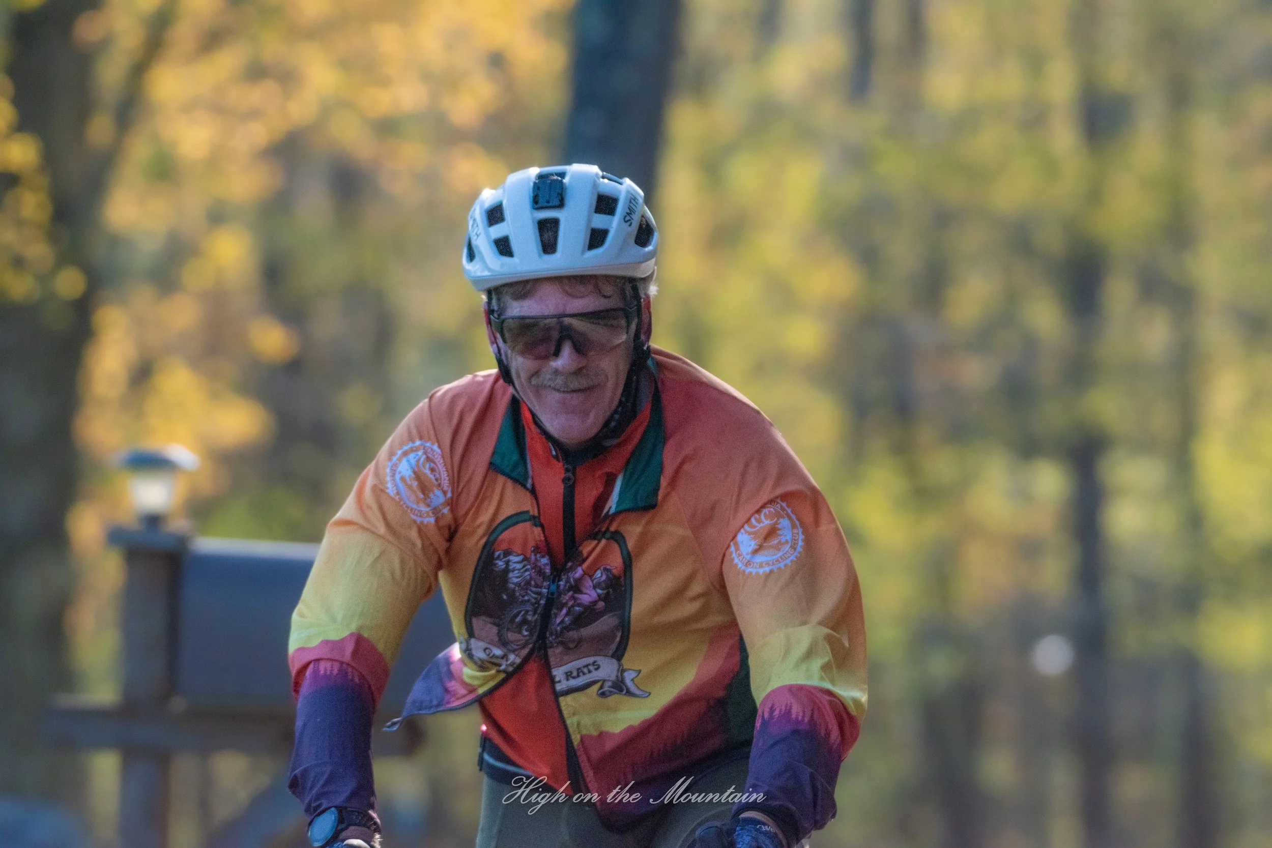 An older man wearing a white bike helmet and sunglasses biking outdoors in a park with trees and sunlight in the background.