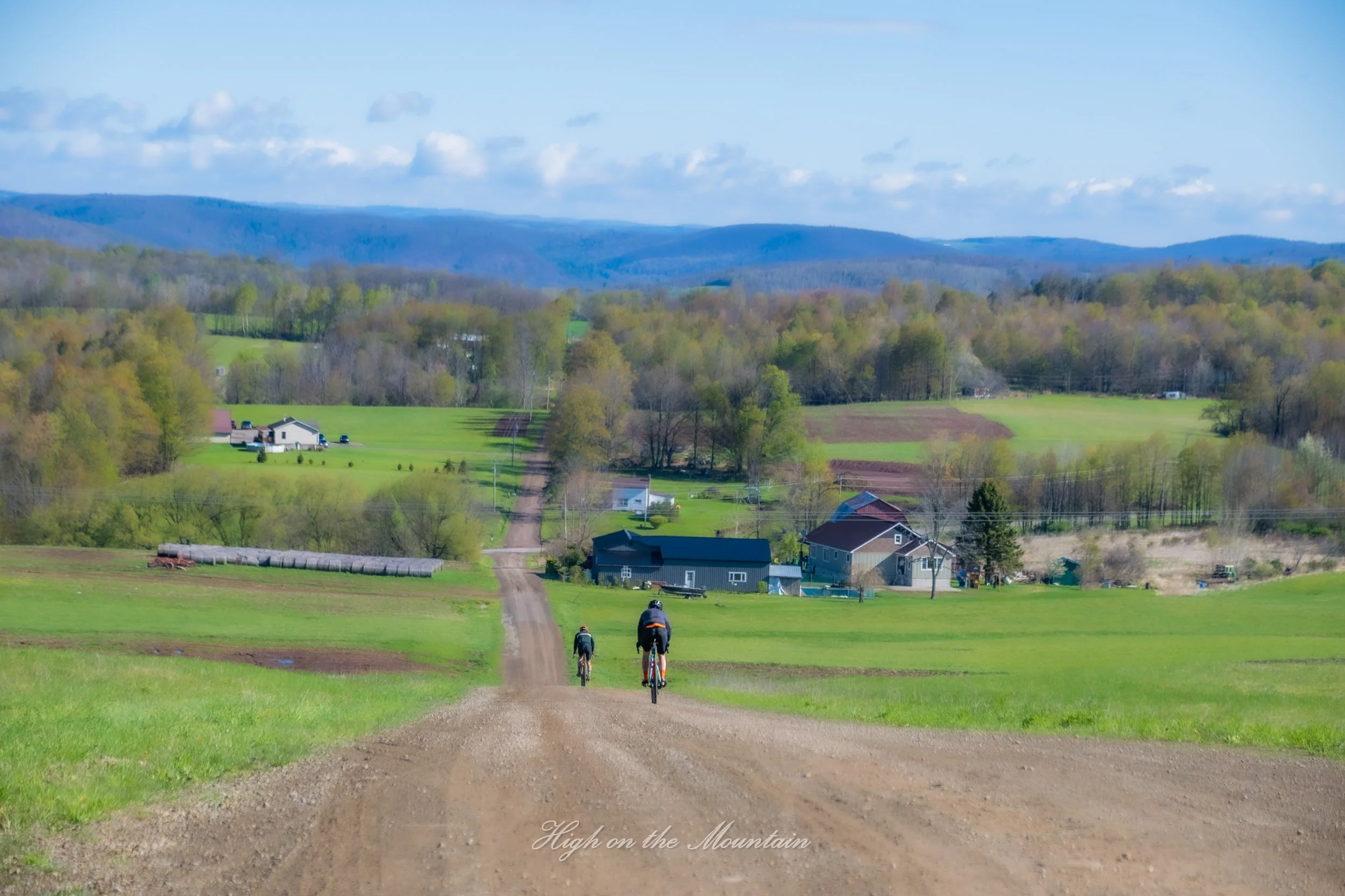 Two people riding bikes on a dirt road through a lush green countryside with houses, trees, and rolling hills under a partly cloudy sky.