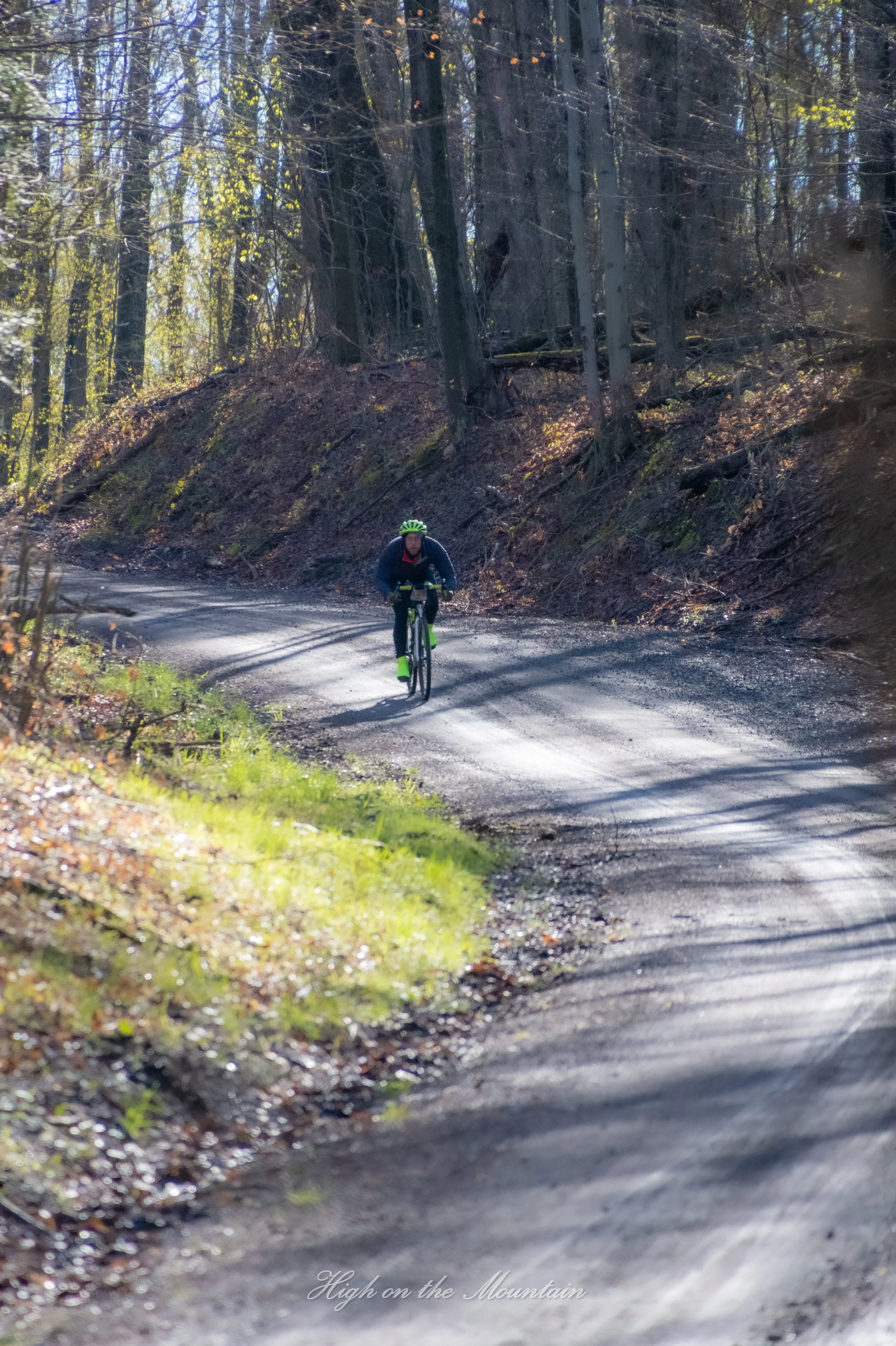 A person riding a bicycle on a dirt trail through a forested area with tall trees and sunlight filtering through the branches. The image has a caption at the bottom that reads 'High on the Mountain.'