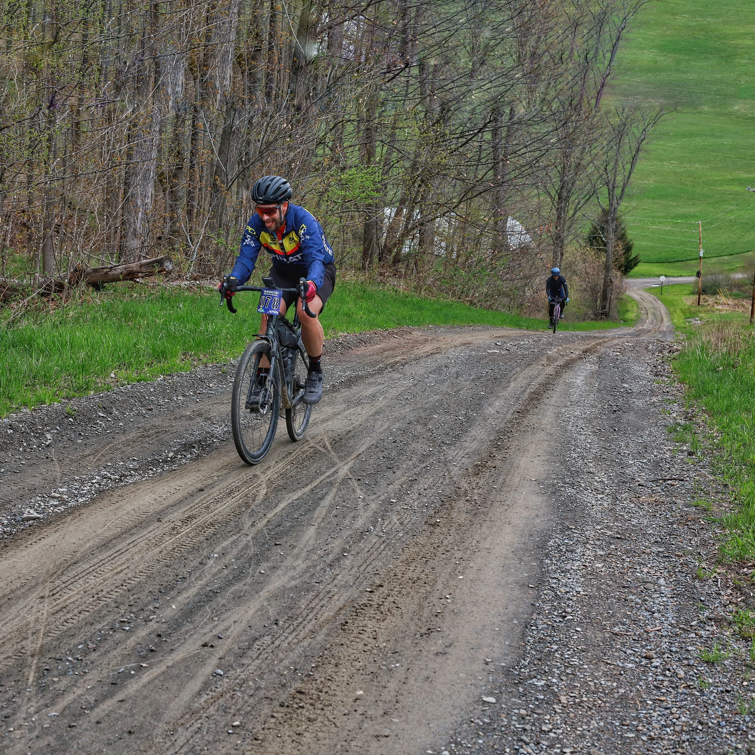 Two cyclists riding on a dirt trail surrounded by grass and trees with green fields in the background.