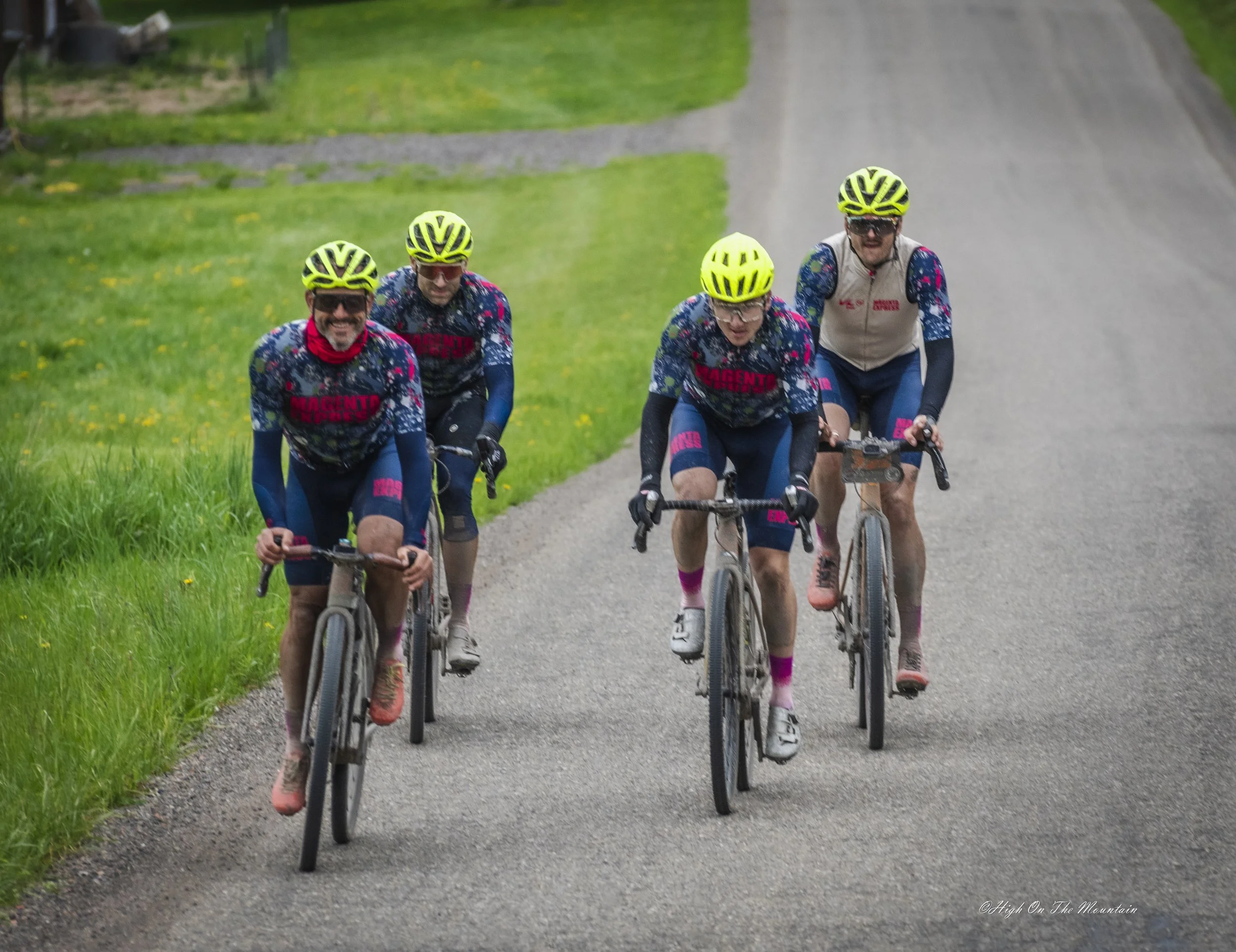 Five cyclists riding mountain bikes on a gravel trail through green fields, wearing helmets and cycling jerseys.