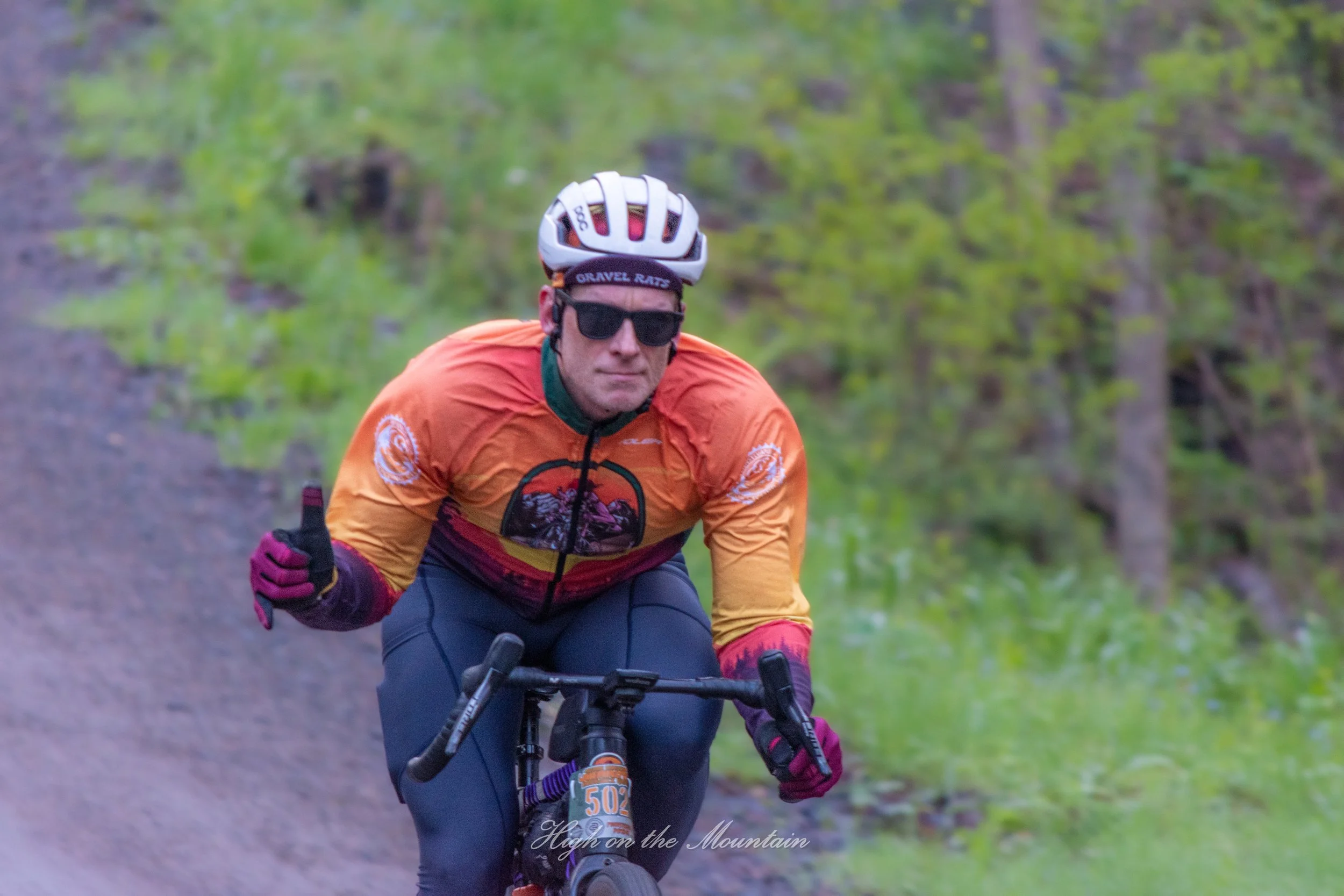 A man riding a mountain bike on a trail surrounded by greenery, wearing an orange jacket, a white helmet, sunglasses, and gloves.