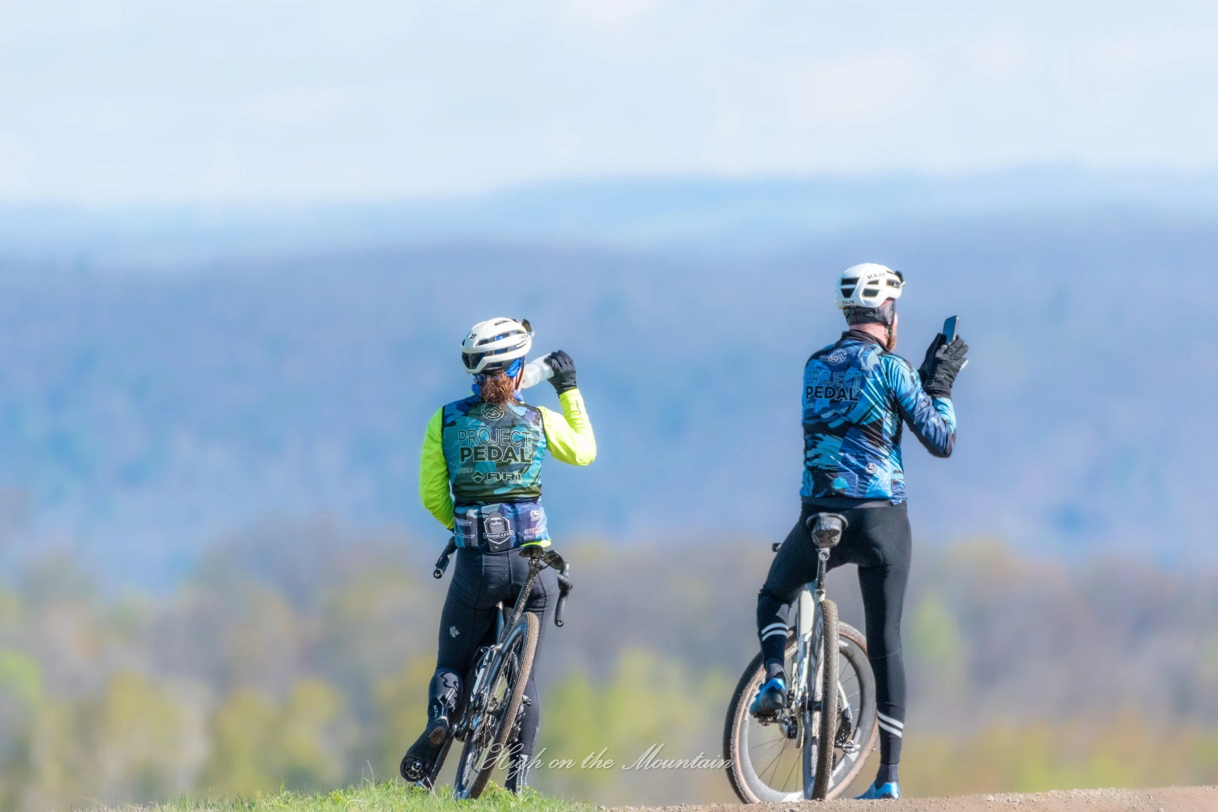 Two cyclists wearing helmets and sports gear, standing with their bikes on a grassy area, overlooking a scenic mountain landscape.