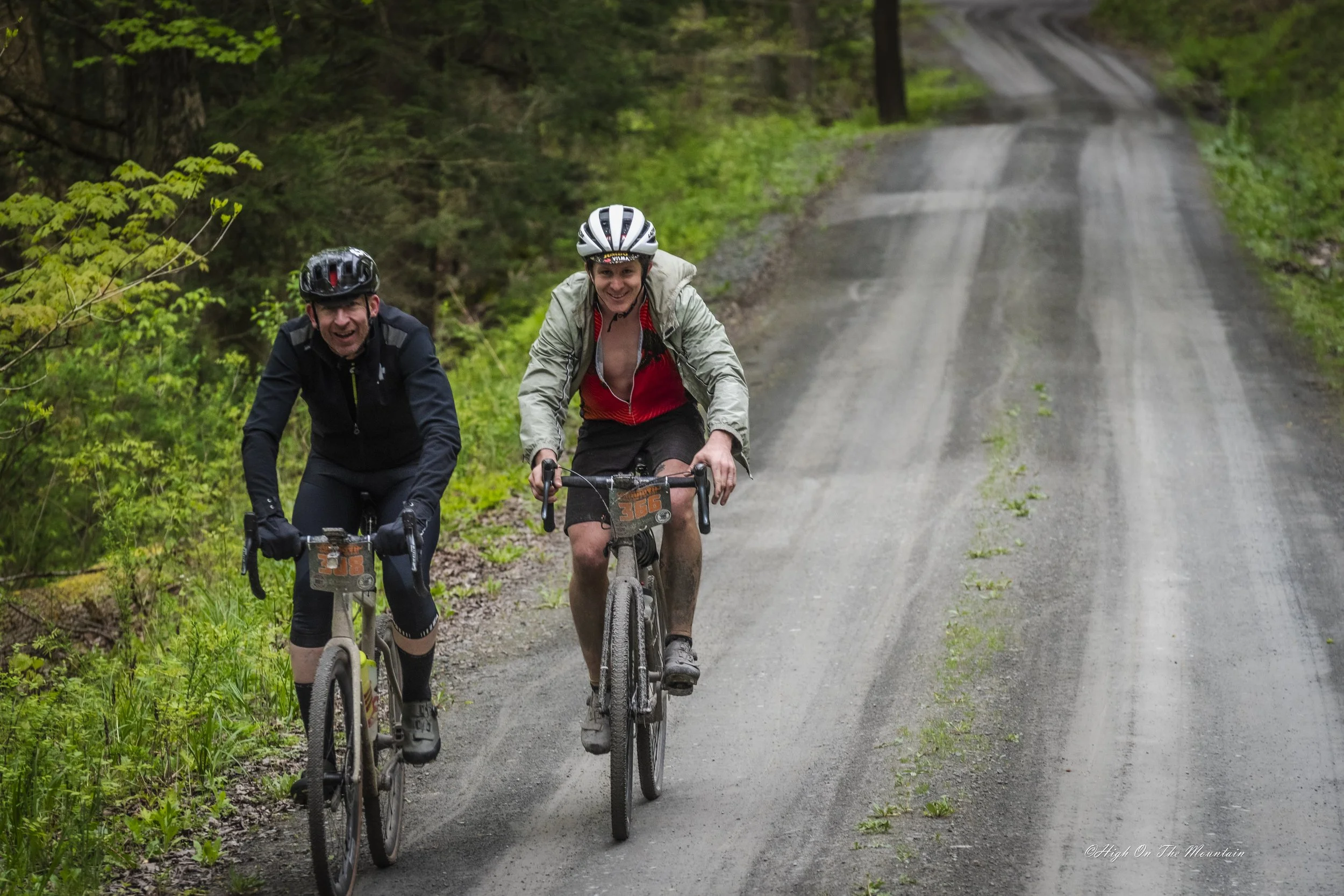 Two men riding mountain bikes on a dirt trail through a green wooded forest, both wearing helmets and smiling.