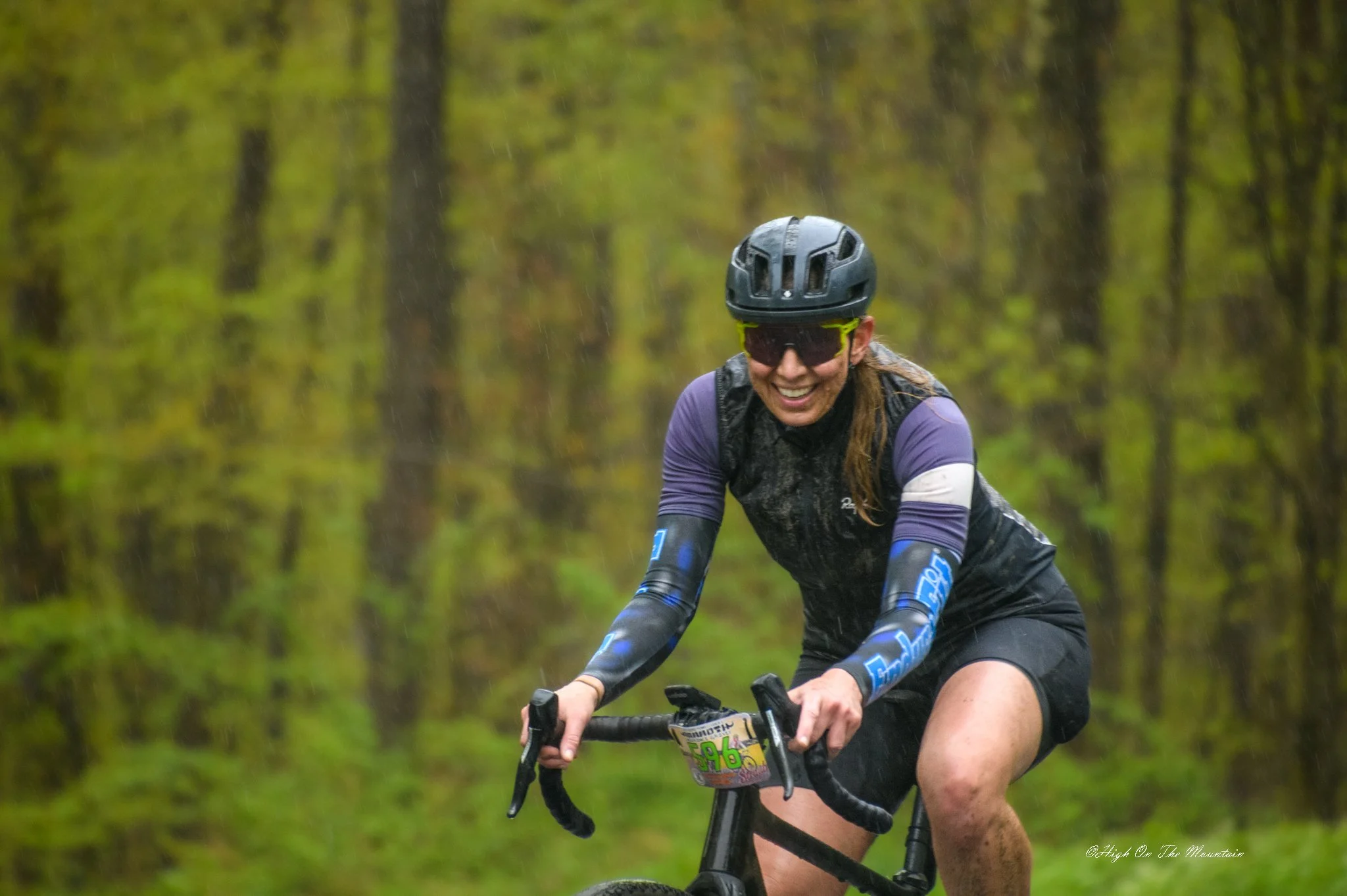 A woman wearing a black helmet, sunglasses, and athletic clothing riding a mountain bike through a rain-soaked forest trail.