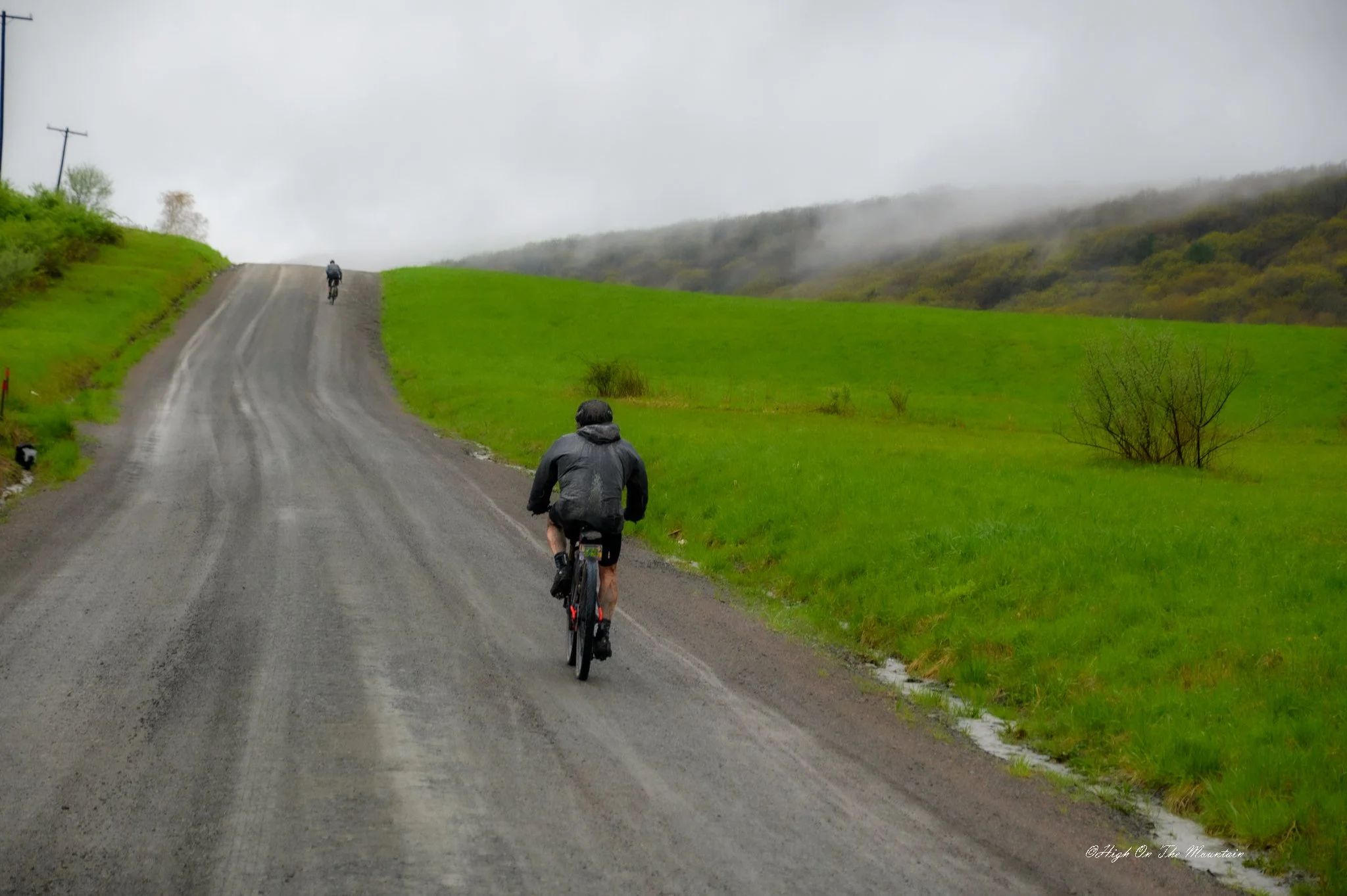 Cyclists riding on a gravel road through green fields on a rainy, foggy day.