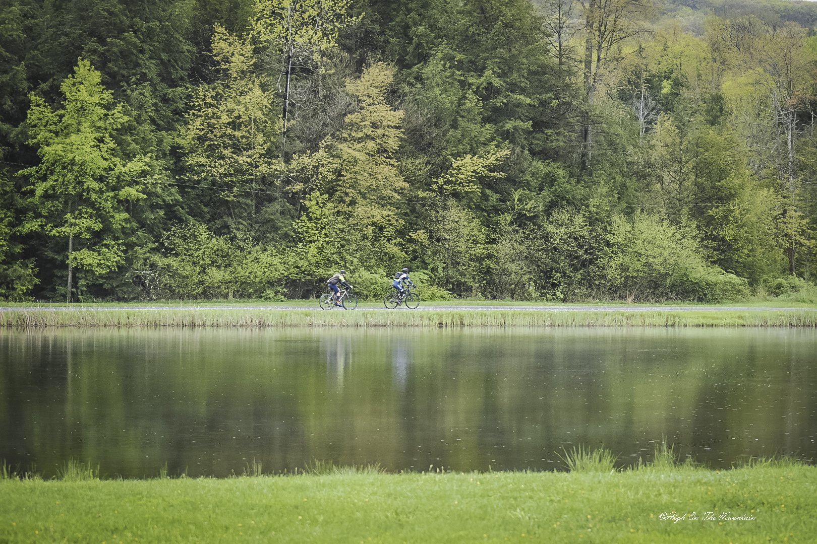 Two cyclists riding along a road beside a body of water, with lush green trees in the background.