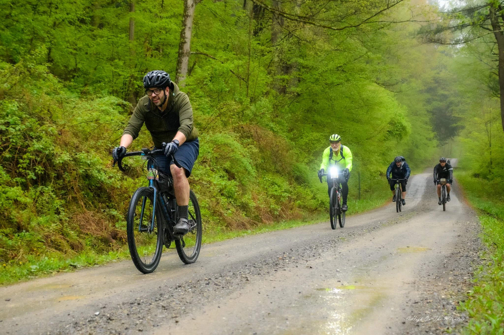 Four cyclists riding on a gravel forest trail with green trees and dense foliage in the background, one cyclist front and center wearing glasses and a helmet, and three further back, one with a bright yellow jacket.