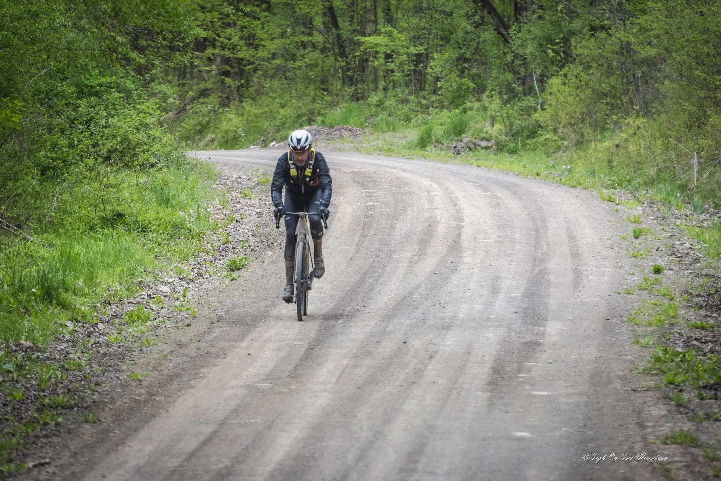 A cyclist wearing a helmet and black outdoor gear riding on a dirt road through a lush green forest.