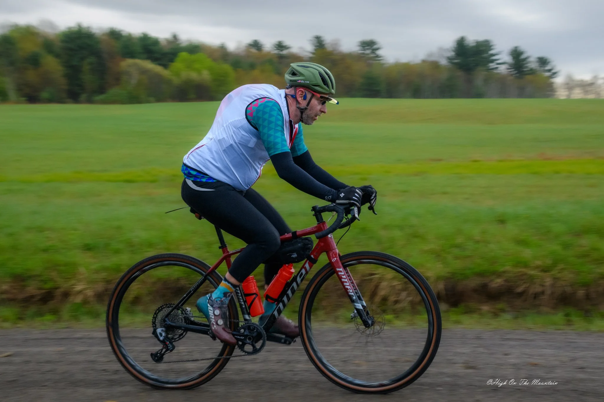 A man wearing a helmet and cycling gear riding a red mountain bike on a trail outdoors, with a grassy field and trees in the background on a cloudy day.