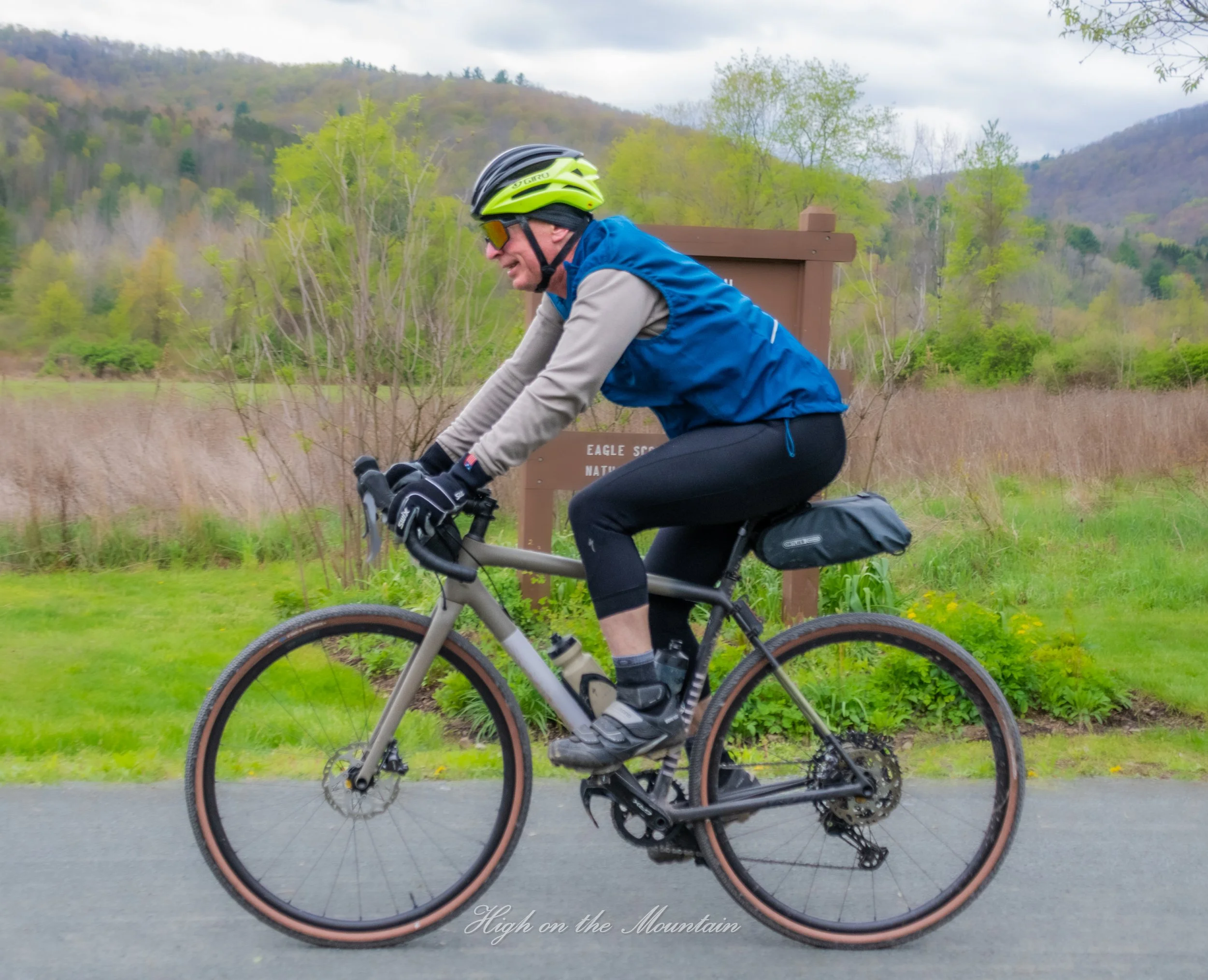 A man riding a bicycle in a rural landscape with green trees, hills, and cloudy sky in the background. The man is wearing a helmet, sunglasses, blue vest, grey long sleeves, and black cycling shorts.