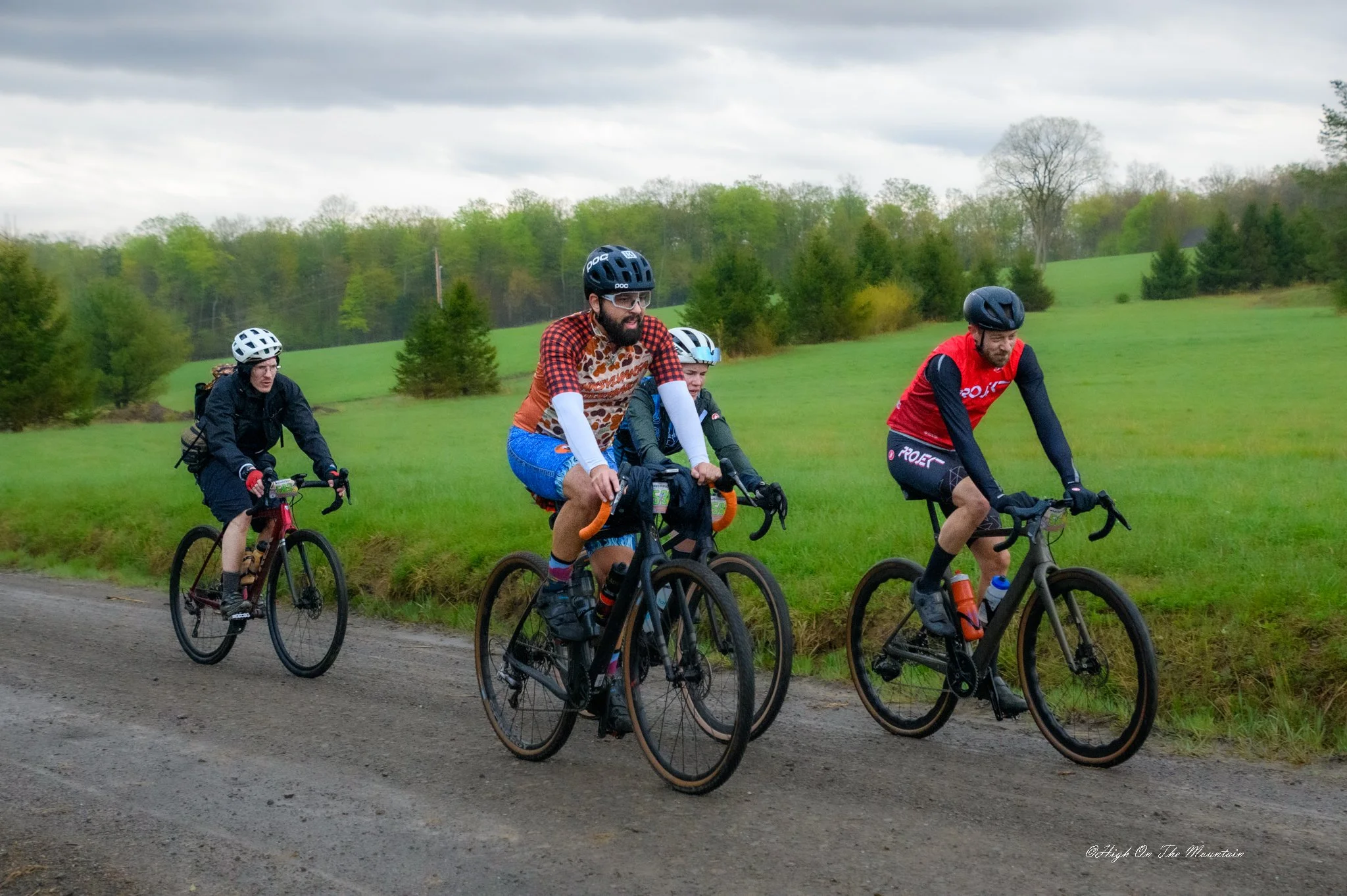 A group of four cyclists riding on a gravel path in a rural area with lush green fields and trees in the background under a cloudy sky.