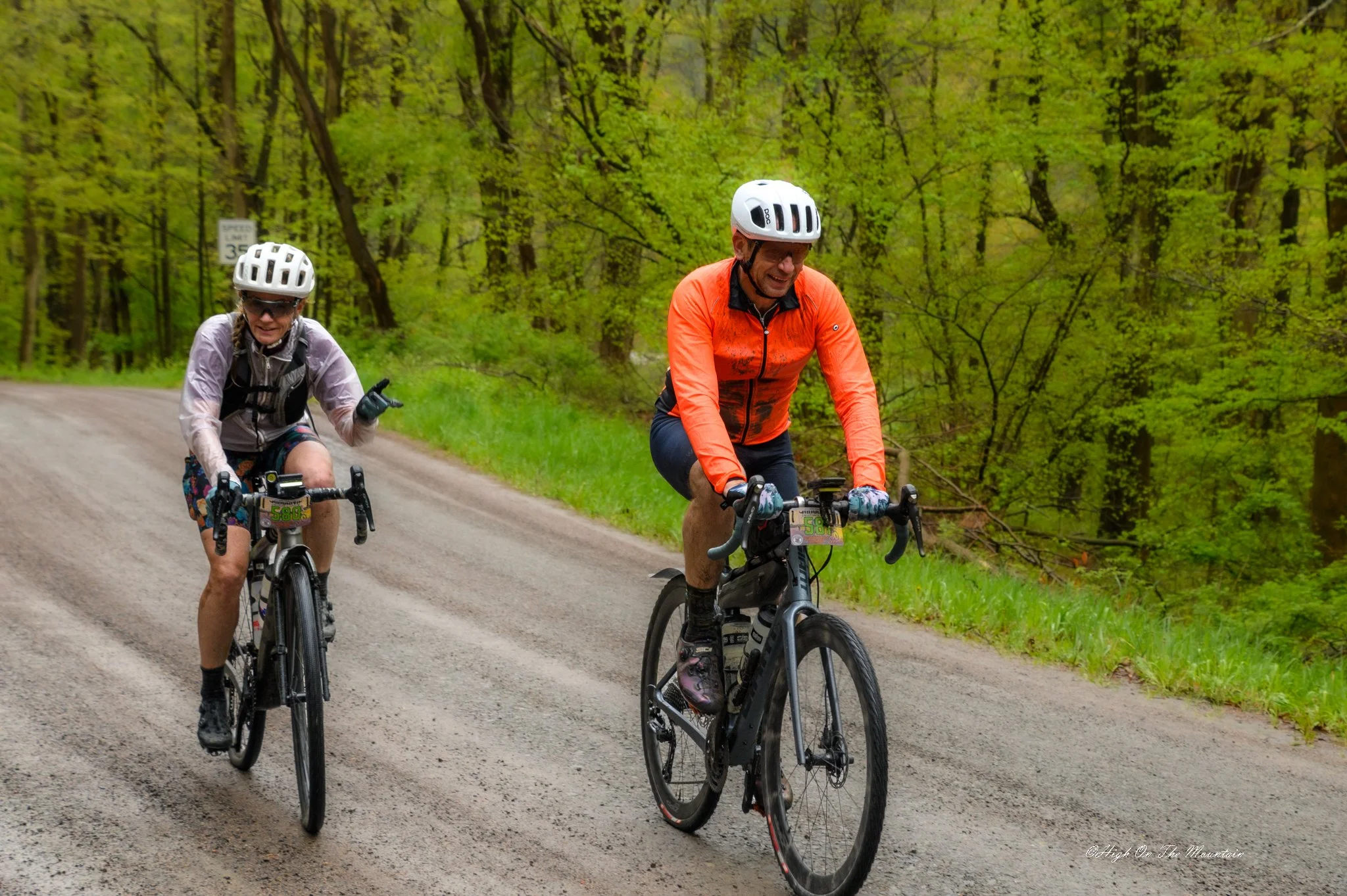 Two people mountain biking on a dirt trail through a green forest, both wearing helmets and athletic gear, smiling and enjoying the ride.