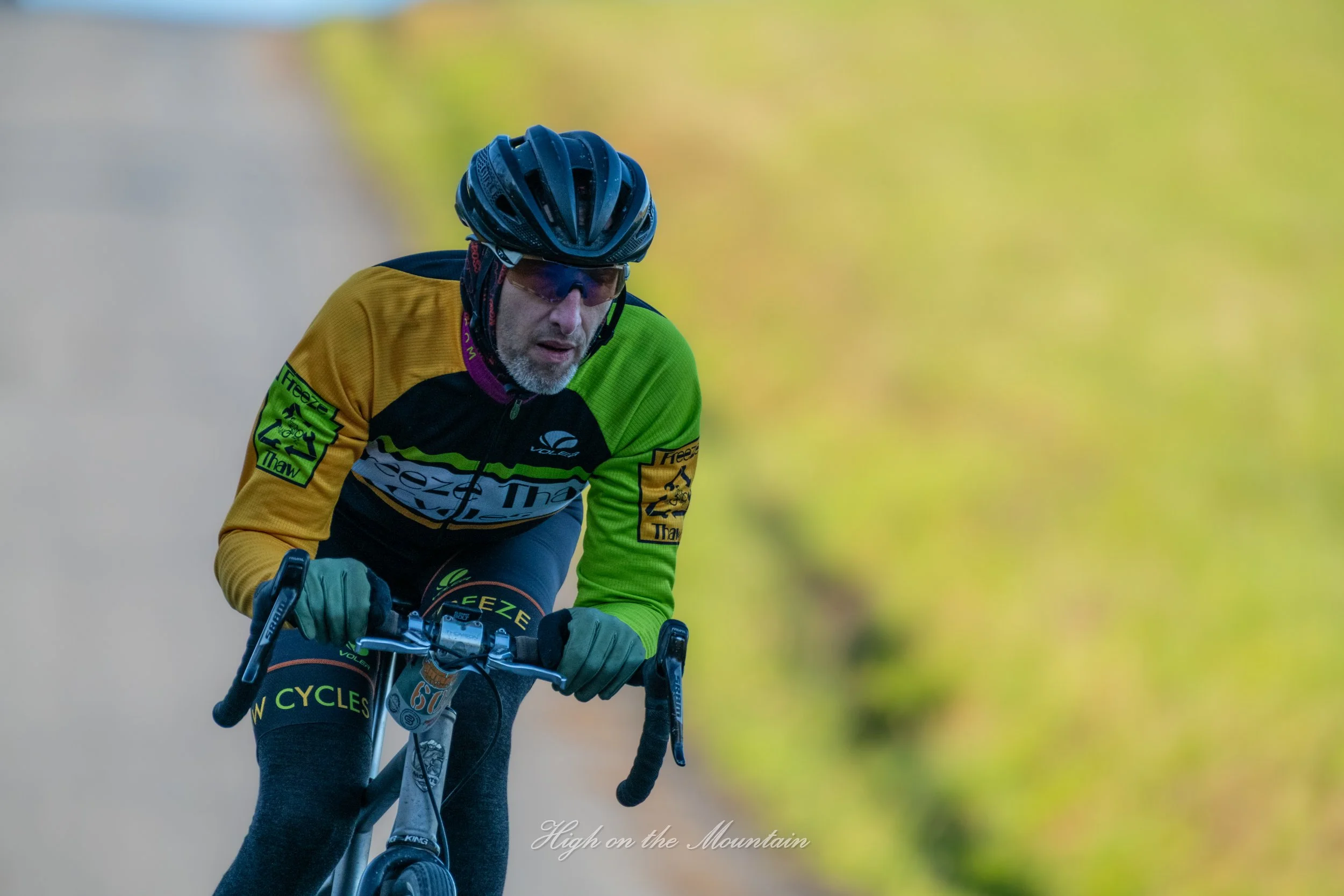 A cyclist wearing a yellow and green jersey, a black helmet, and sunglasses rides a mountain bike on a trail with blurred greenery in the background.
