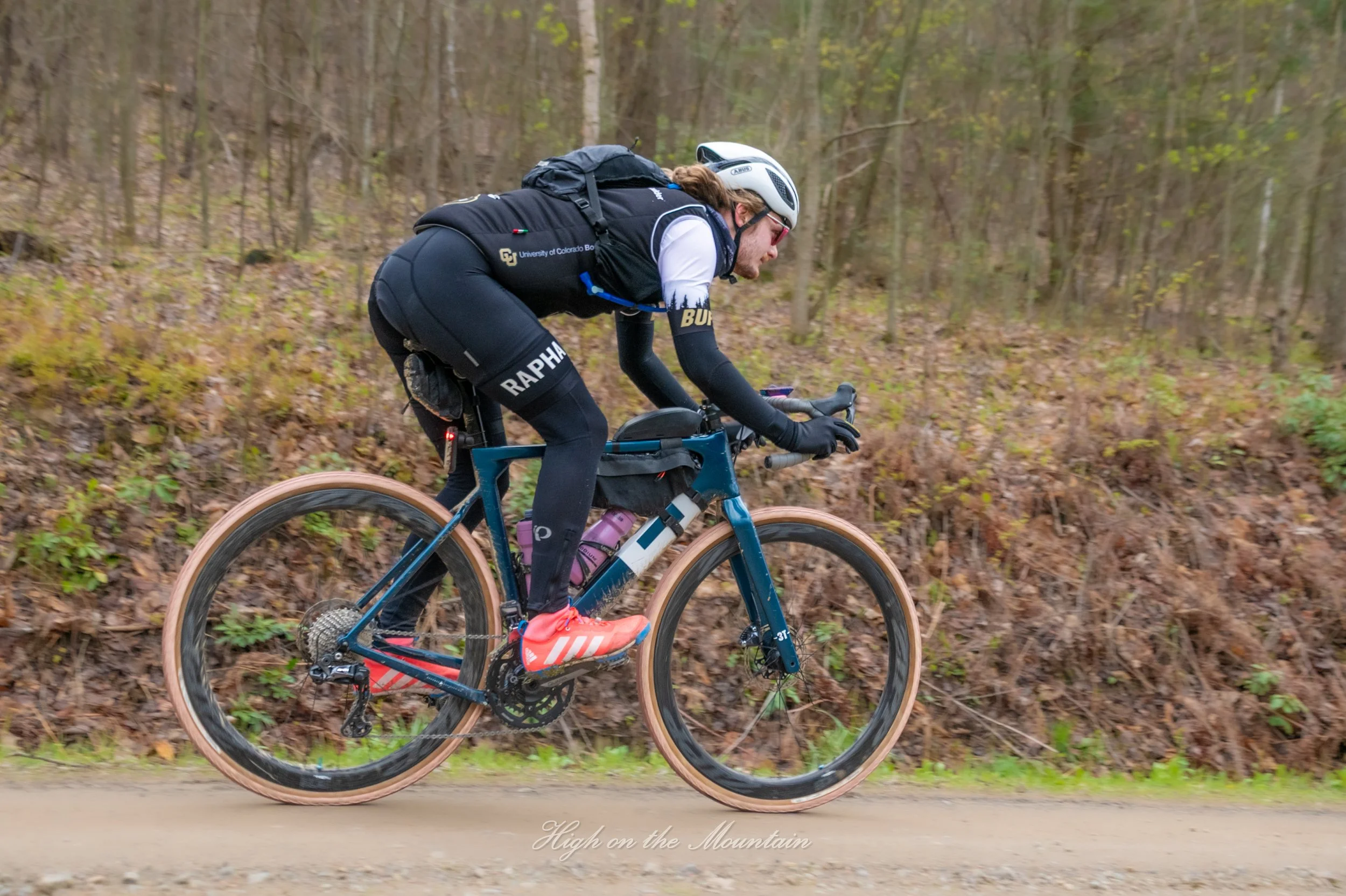 A person riding a mountain bike on a trail through a forested area with trees and fallen leaves, wearing a helmet, sunglasses, a backpack, and athletic clothing with branding.