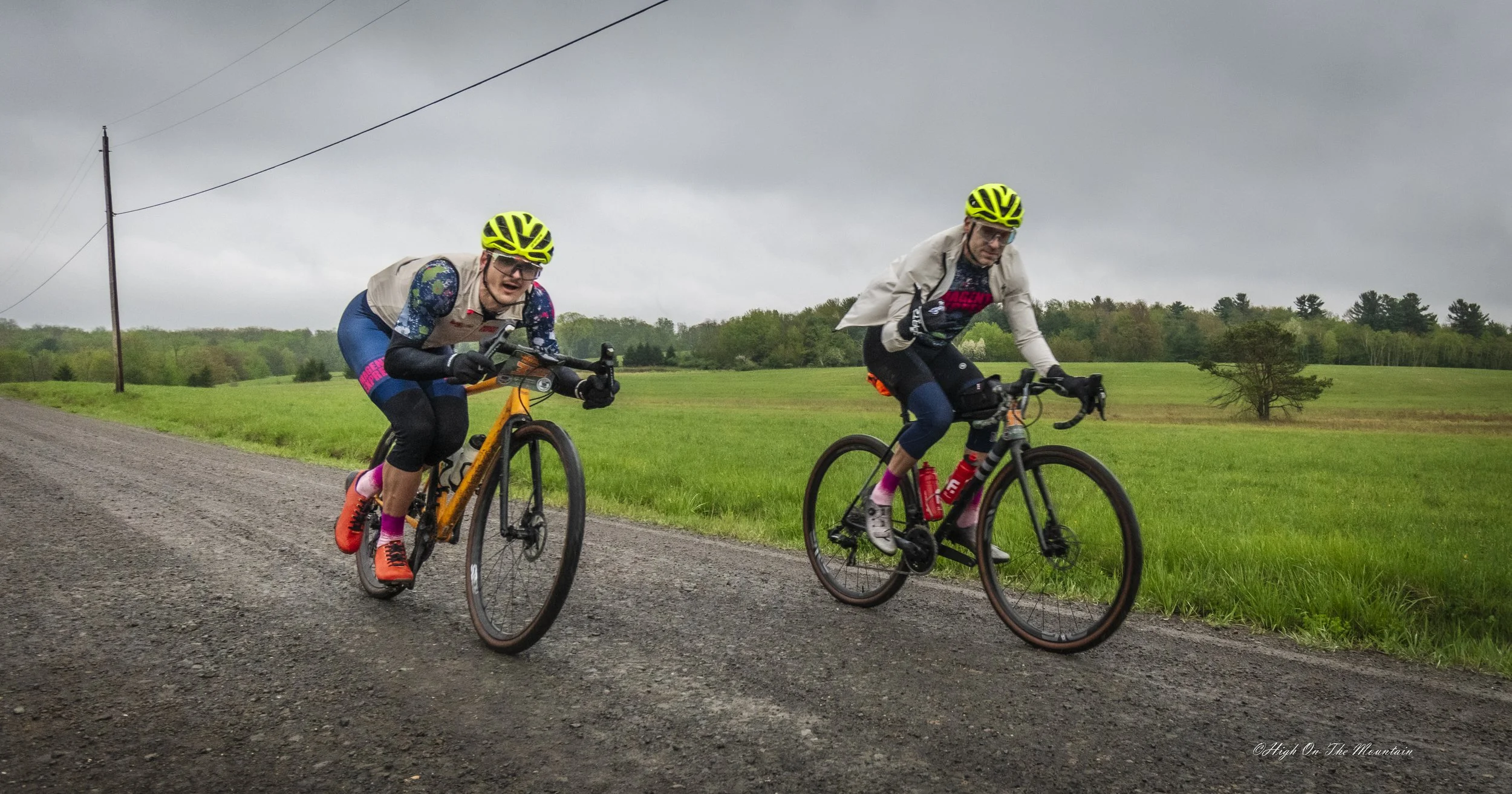 Two women riding bicycles on a gravel road in a rural area with green fields and trees under an overcast sky.