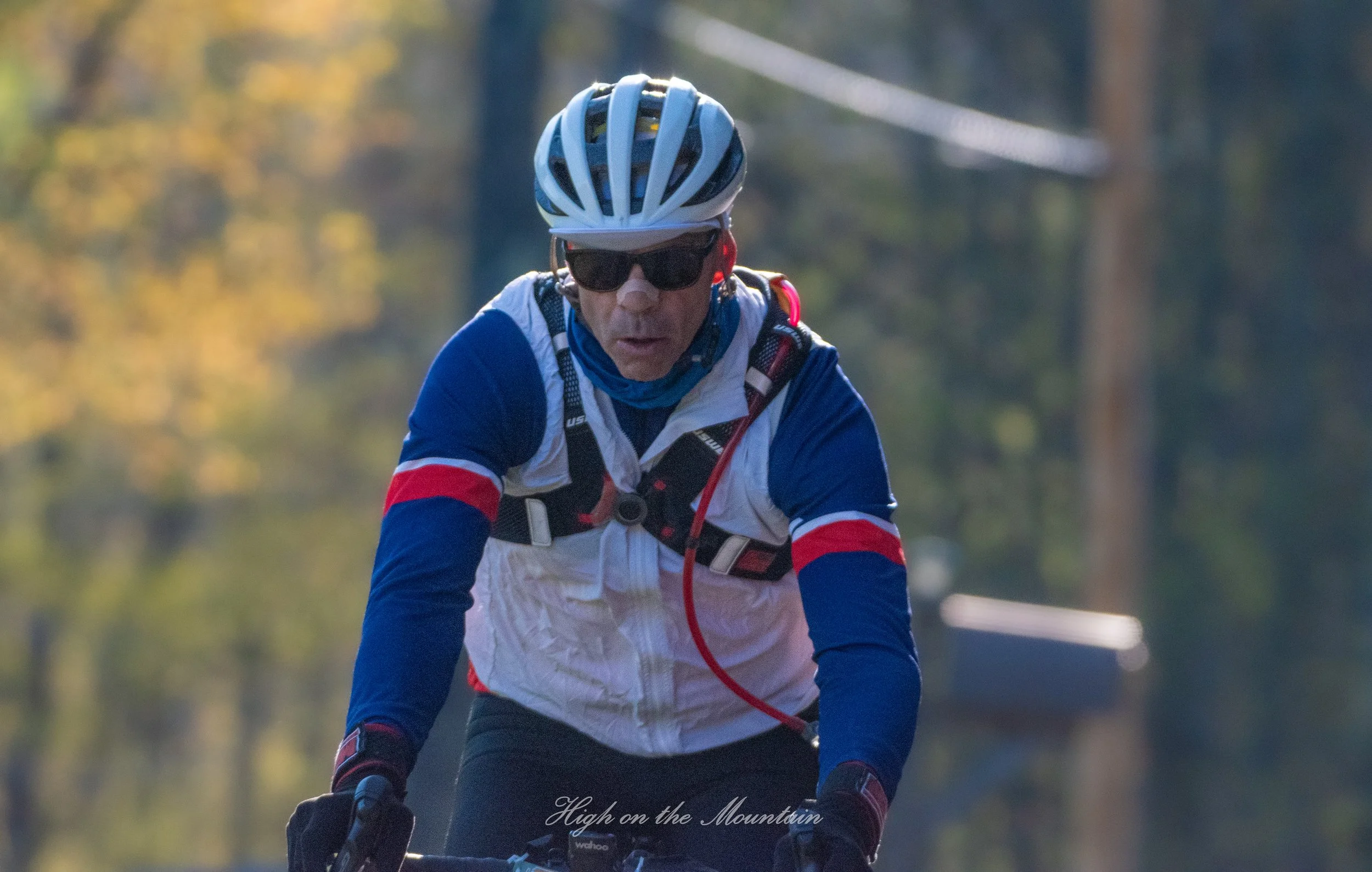 A man riding a mountain bike outdoors wearing a white and blue cycling jersey, a helmet, sunglasses, and a neck gaiter, with trees and a bridge in the background.