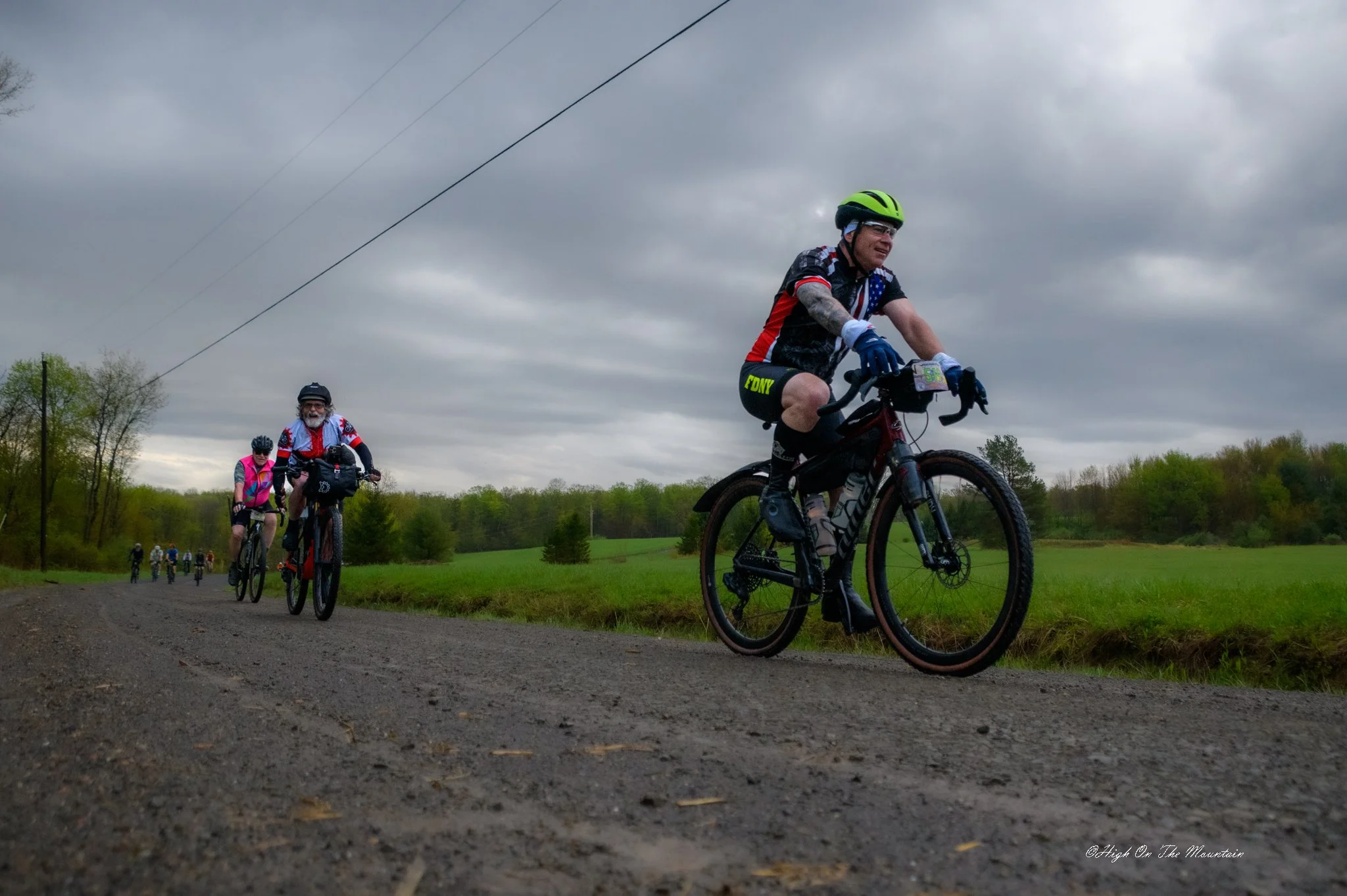 A group of cyclists riding on a dirt trail in a rural area on a cloudy day, with trees and green fields in the background.