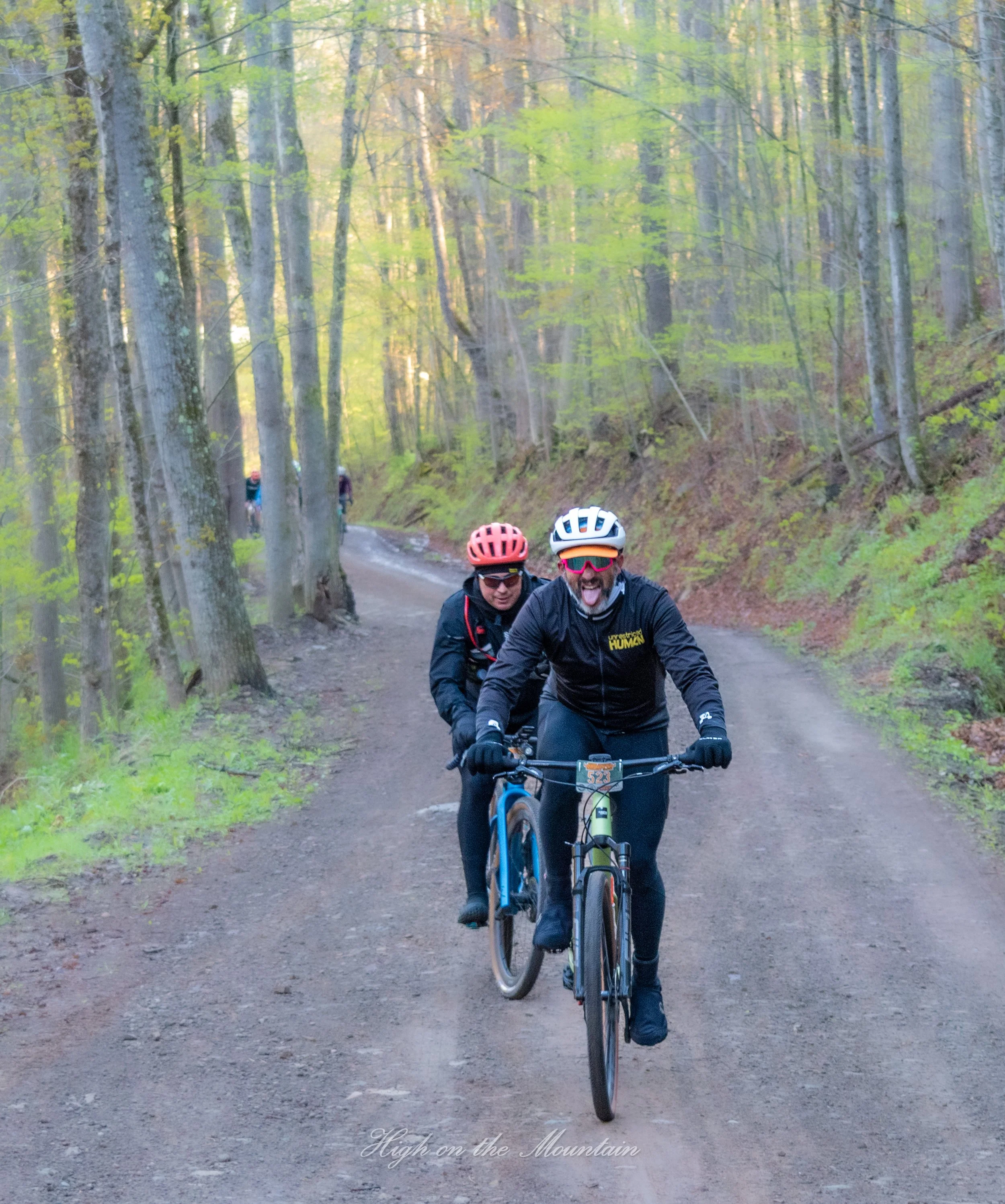 Two mountain bikers riding on a forest trail with green trees and sunlight filtering through. The bikers are wearing helmets and sunglasses, and the person in front is sticking out their tongue. The background shows more bikers on the trail among den